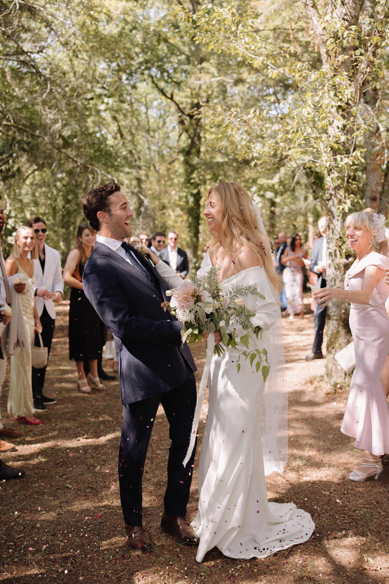 Couple in confetti send-off on wooded path bride in off-shoulder lace gown with blush dahlia bouquet guests throwing petals