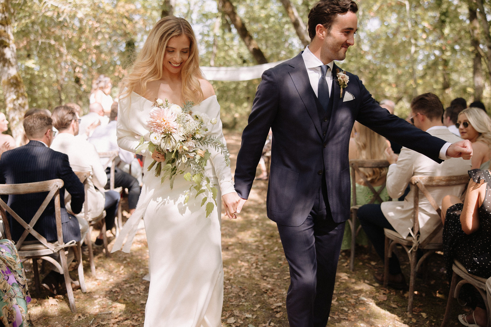 Couple walking back down woodland aisle after ceremony with blush dahlia bouquet