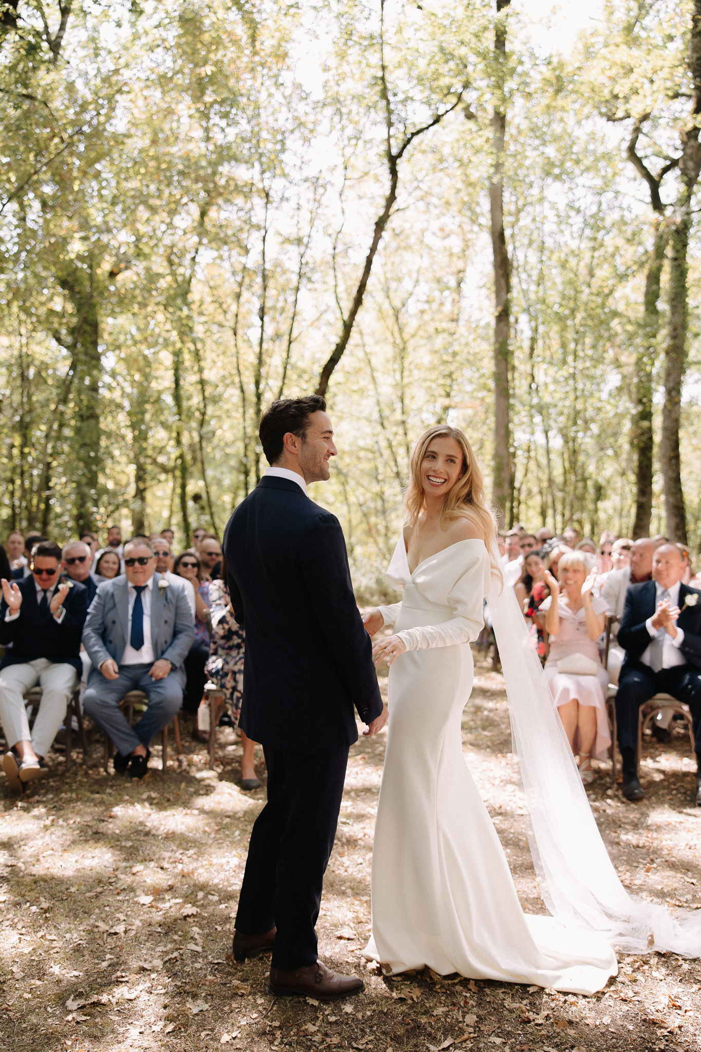 Bride laughing and holding grooms hand after woodland ceremony with seated guests applauding in background