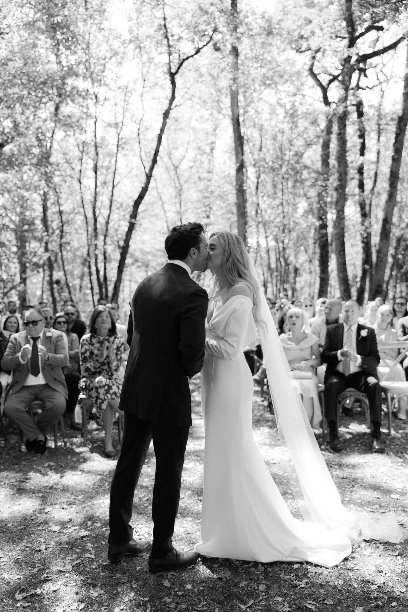 Black and white first kiss from behind in woodland ceremony with thirty guests and dappled forest light