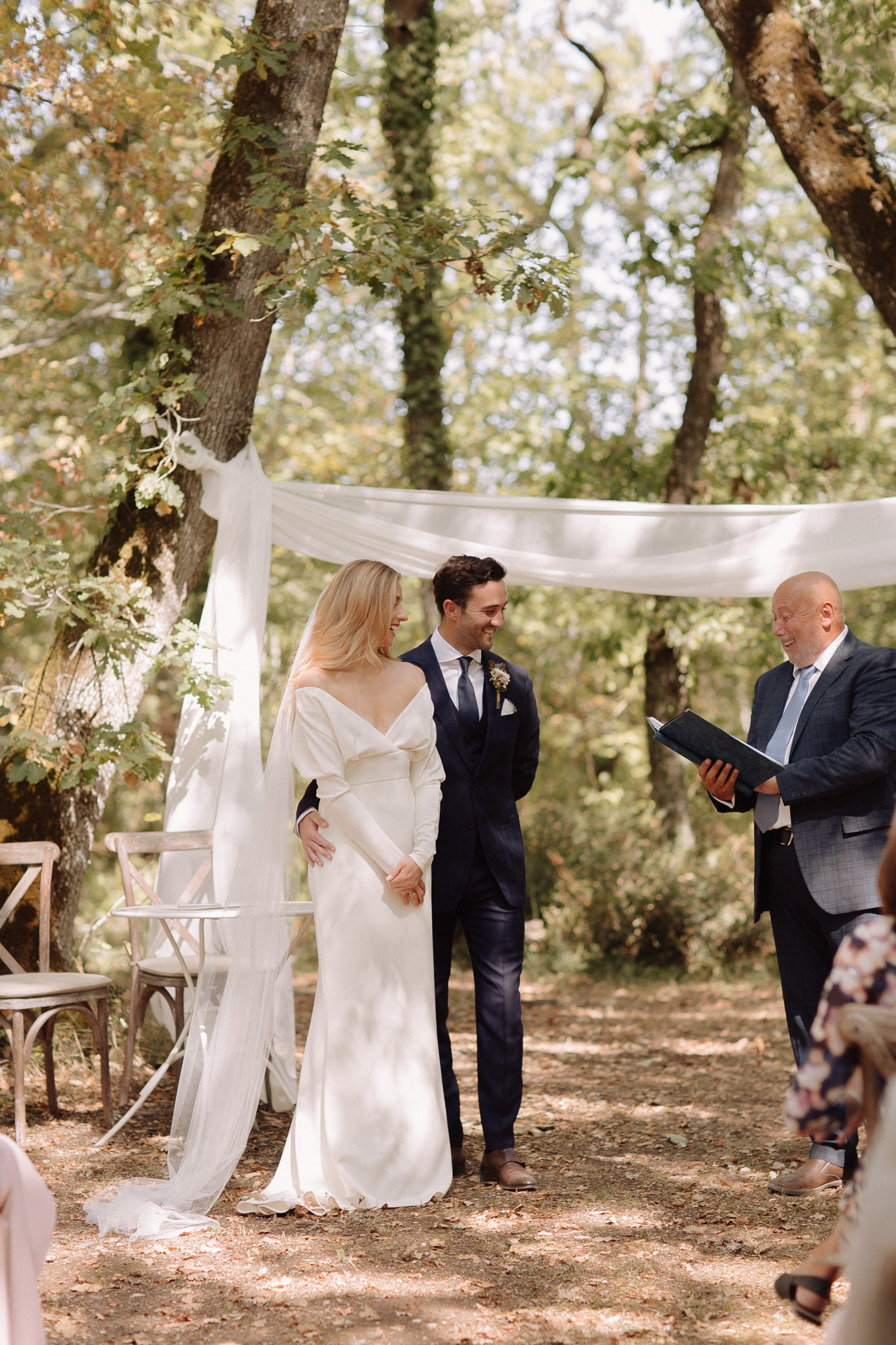 Couple under white draped fabric arch in woodland ceremony bride in ivory wrap gown officiant with ceremony book