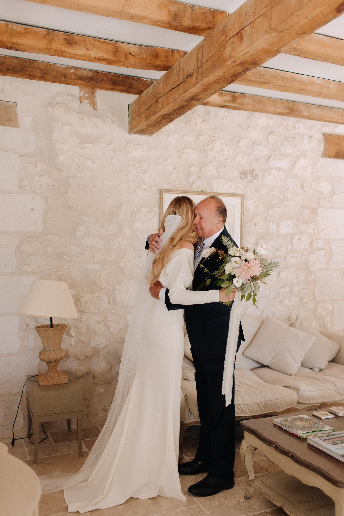 Bride in ivory gown embracing father holding white dahlia and fern bouquet in rustic stone room