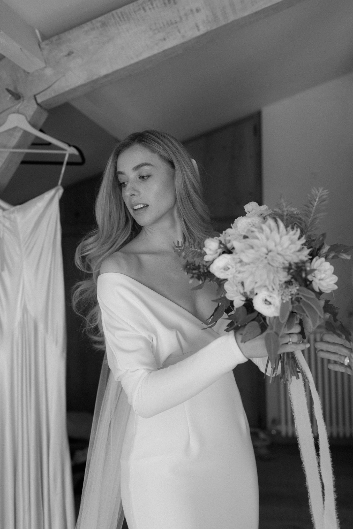 Black-and-white portrait of bride in off-shoulder dress holding a dahlia and ranunculus bouquet with trailing ribbons