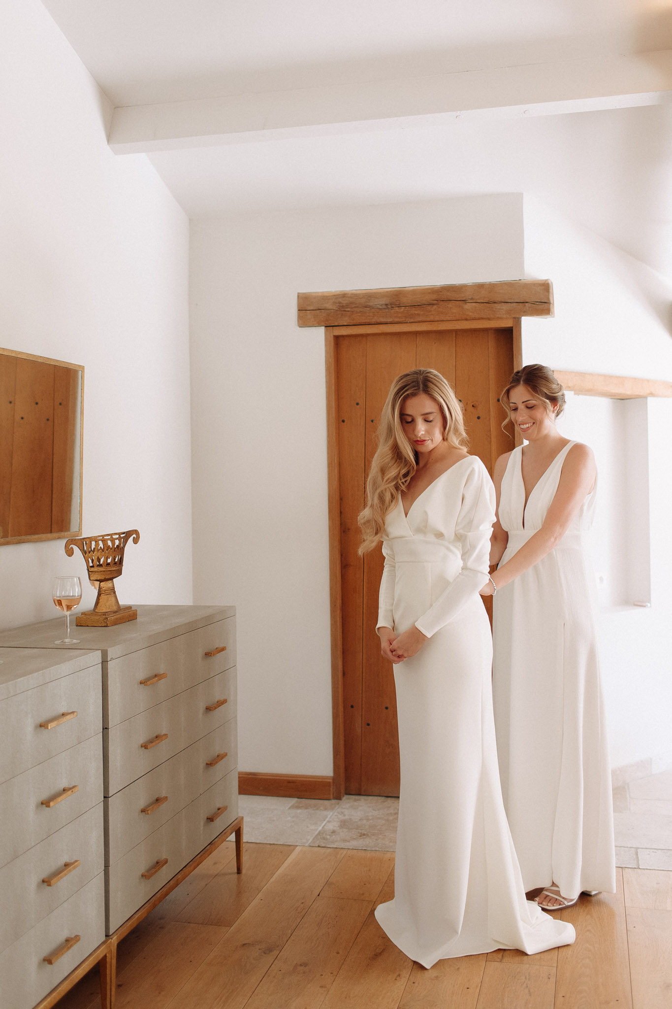 A getting-ready scene captured indoors in a bright, whitewashed room with exposed wooden ceiling beams, wide-plank oak flooring, and a natural wood-framed door. The bride, a blonde woman with loose waves, wears a fitted ivory long-sleeve gown with a deep V-neckline and sleek silhouette, and looks downward as a second woman — likely a bridesmaid or maid of honor — stands behind her smiling and fastening or adjusting the back of the dress. The second woman wears a white sleeveless deep V-neck floor-length gown with strappy sandals. A grey dresser with gold hardware sits to the left, topped with a gold ornate urn and a glass of rosé wine, alongside a gold-framed mirror. The overall styling is modern and minimal, with a warm neutral palette throughout the room. Full-length portrait shot.