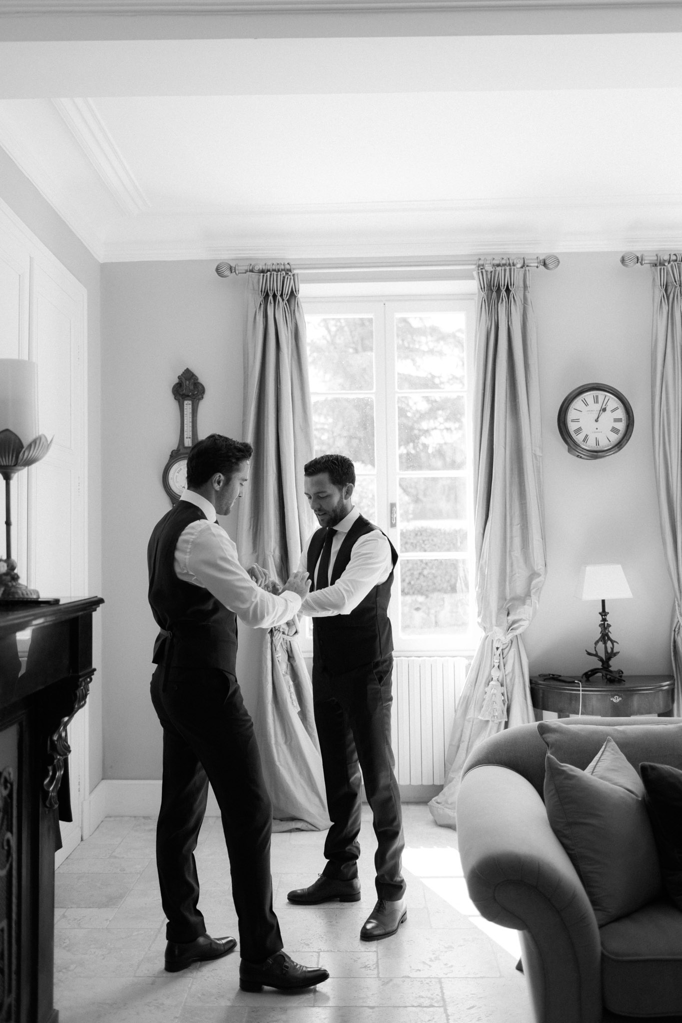 Black and white groomsman helping groom with tie in chateau room with carved fireplace and tall window