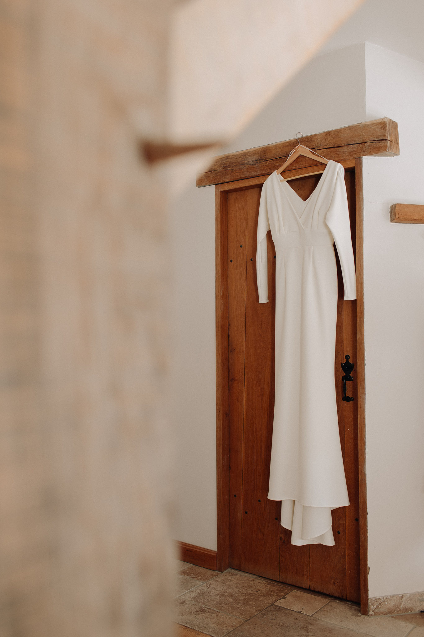 A detail shot of an ivory, long-sleeved wedding dress hanging on a wooden hanger from a rustic wooden door frame with an exposed beam lintel. The dress features a V-neckline, fitted long sleeves, a clean A-line silhouette, and a small train pooling at the bottom — a minimal, modern design with no embellishment. The door is a warm-toned wood with black iron hardware, set against white-plastered walls and stone tile flooring, consistent with a French farmhouse or mas interior. The foreground is intentionally blurred, and the composition is a medium shot framing the dress centrally against the door.