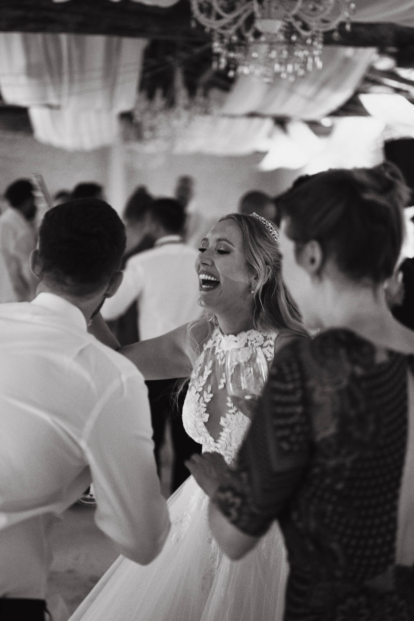 This black-and-white image captures the bride laughing openly on the dance floor during the wedding reception, surrounded by guests. She is wearing a halter-neck lace gown with floral appliqué detailing and a delicate tiara headpiece, and is holding a wine glass. The setting appears to be under a draped tent or marquee with crystal chandeliers visible overhead, creating a classic reception atmosphere. The image is a mid-shot taken at a slight angle, with the background guests rendered soft and out of focus, drawing attention to the bride's expression at center frame.