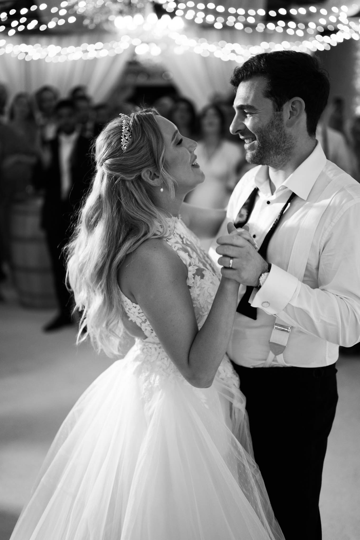 This black-and-white image captures the couple's first dance at an indoor reception, with guests visible watching in the soft-focus background. The bride wears a lace-bodice ballgown with a low open back and a full tulle skirt, accessorized with a jeweled tiara headpiece and pearl drop earrings; the groom is in a white dress shirt with dark trousers and a loosened tie, his jacket removed. Both are laughing and facing each other, their hands clasped between them, conveying a candid, joyful moment. Overhead, dense fairy lights and draped white fabric create a warm, glowing canopy above the dance floor, giving the background strong bokeh contrast against the sharp foreground tones.