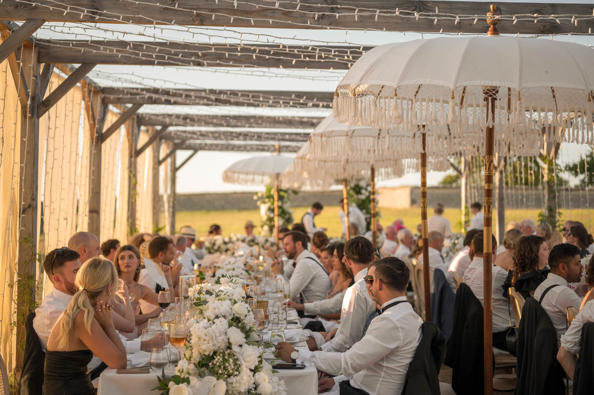 Guests seated at long banquet tables under fairy-lit wooden pergola with white rose runners and parasols