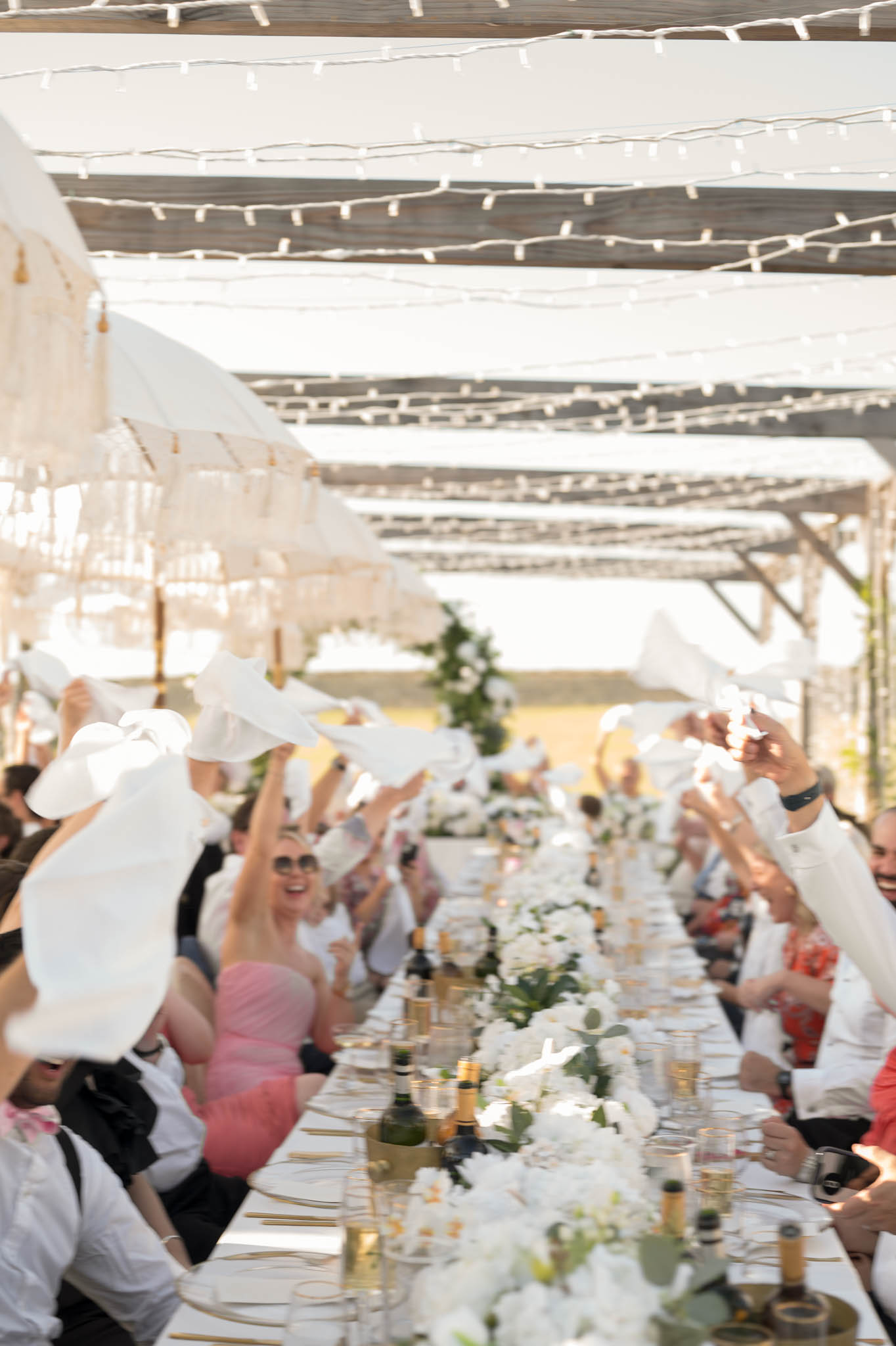 Guests waving white napkins at pergola dinner table with hydrangea runner and fairy lights overhead