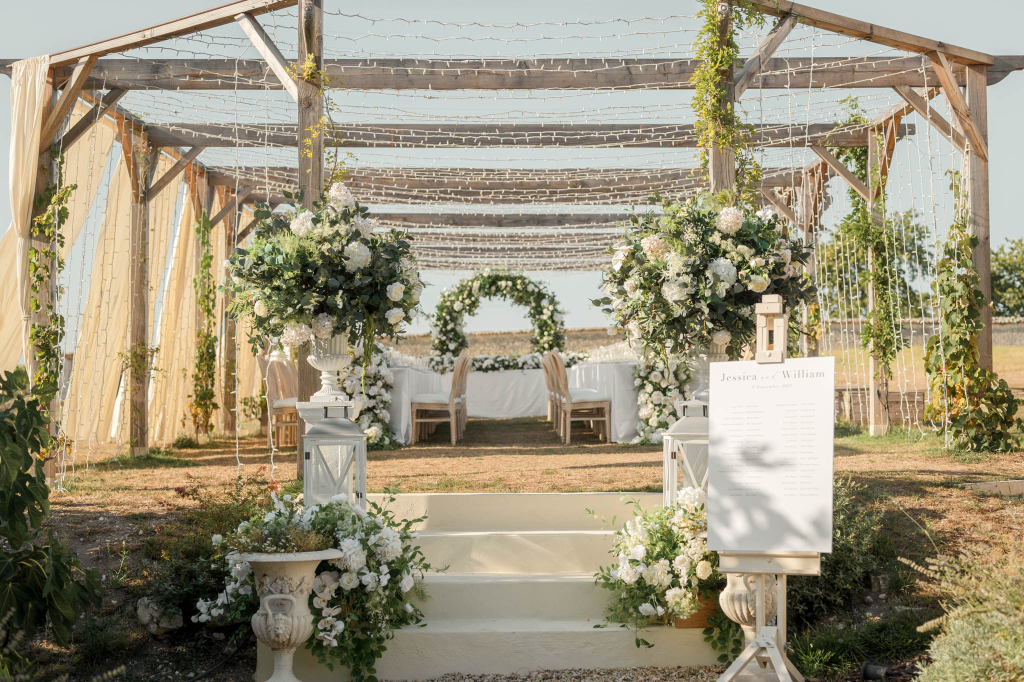 Outdoor ceremony pergola with fairy lights and white hydrangea urns flanking steps leading to a circular greenery arch altar