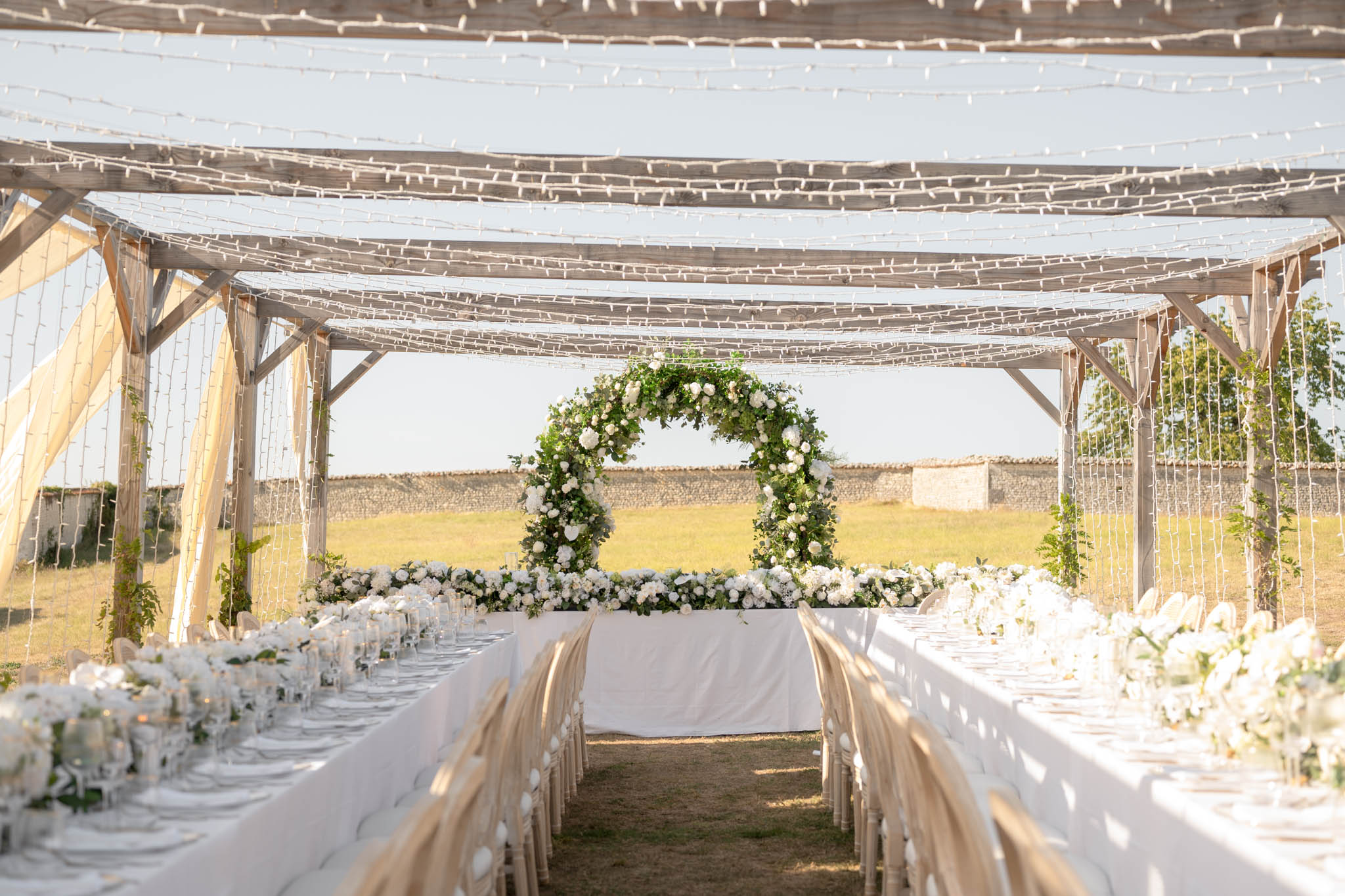 Pergola reception with fairy lights, white rose centerpieces, circular floral arch, and sheer ivory drapes