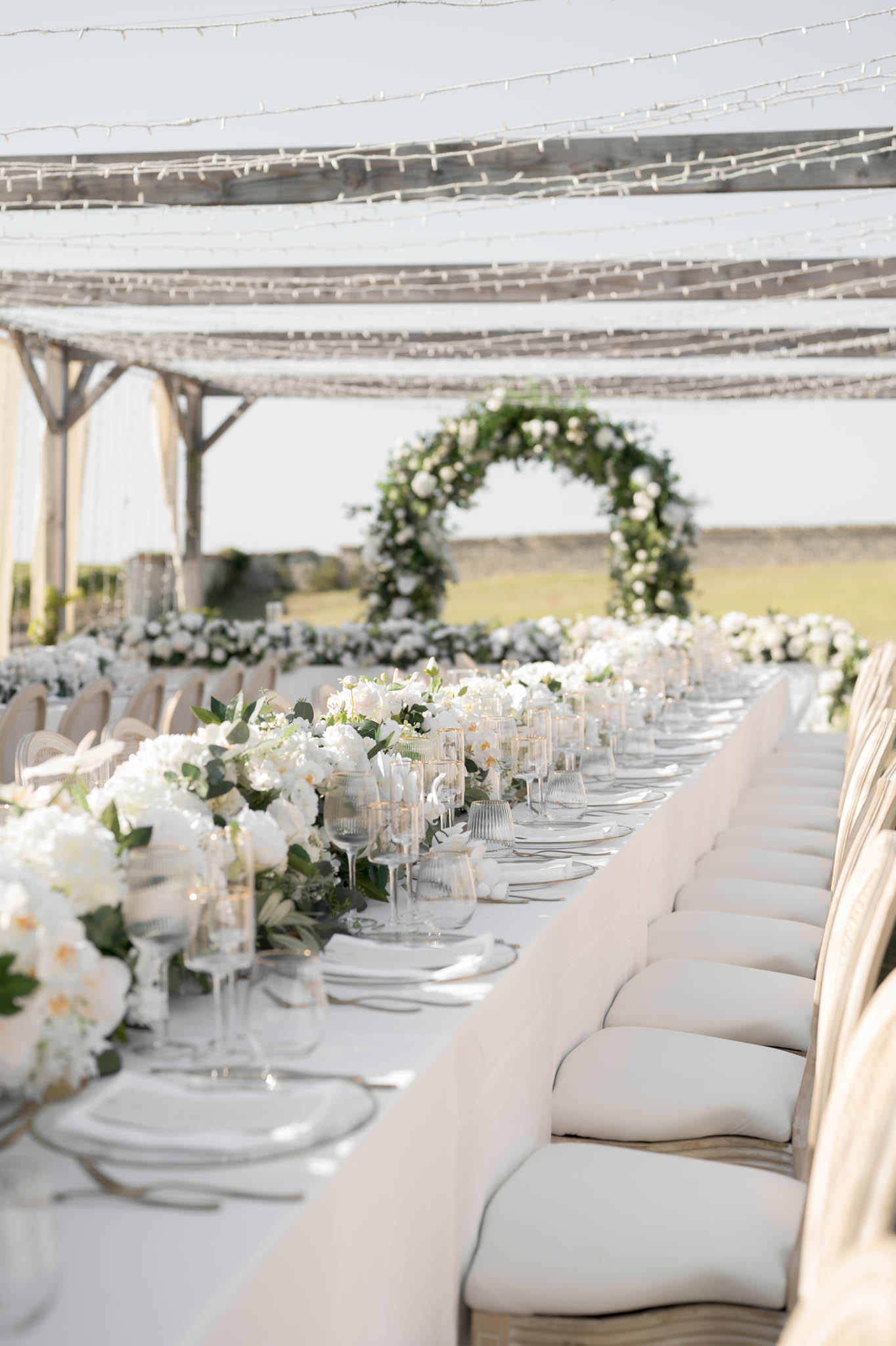 Reception table with white peony and orchid runner and circular floral arch under pergola