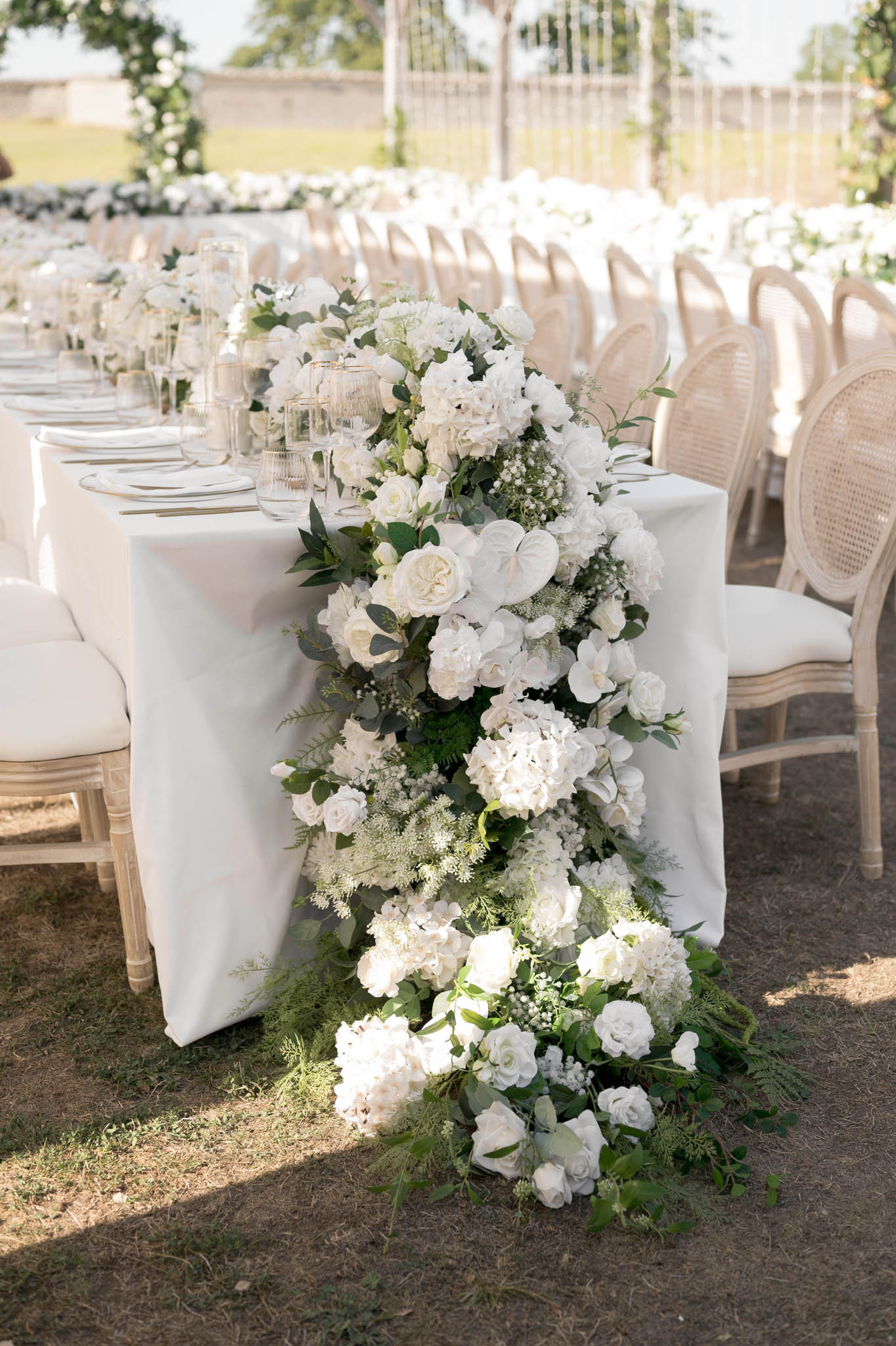 Close-up detail shot of an outdoor wedding reception table setup, focusing on a large cascading floral arrangement at the end of a long banquet table covered in a white linen cloth. The arrangement is composed entirely of white flowers — including garden roses, hydrangeas, orchids, white anthurium, baby's breath, and lisianthus — mixed with lush green eucalyptus, fern fronds, and trailing foliage that spills down the side of the table and continues onto the ground below. The table is set with white plates, gold-rimmed glassware, and ribbed glass candle holders running along its length. Seating consists of natural wood Louis XVI-style cane-back chairs with ivory cushions, positioned on both sides of the table. In the background, additional long reception tables are visible, along with a hanging installation featuring white floral and crystal or ribbon details, suggesting a fully all-white and green decor palette with a classic, garden-style aesthetic.