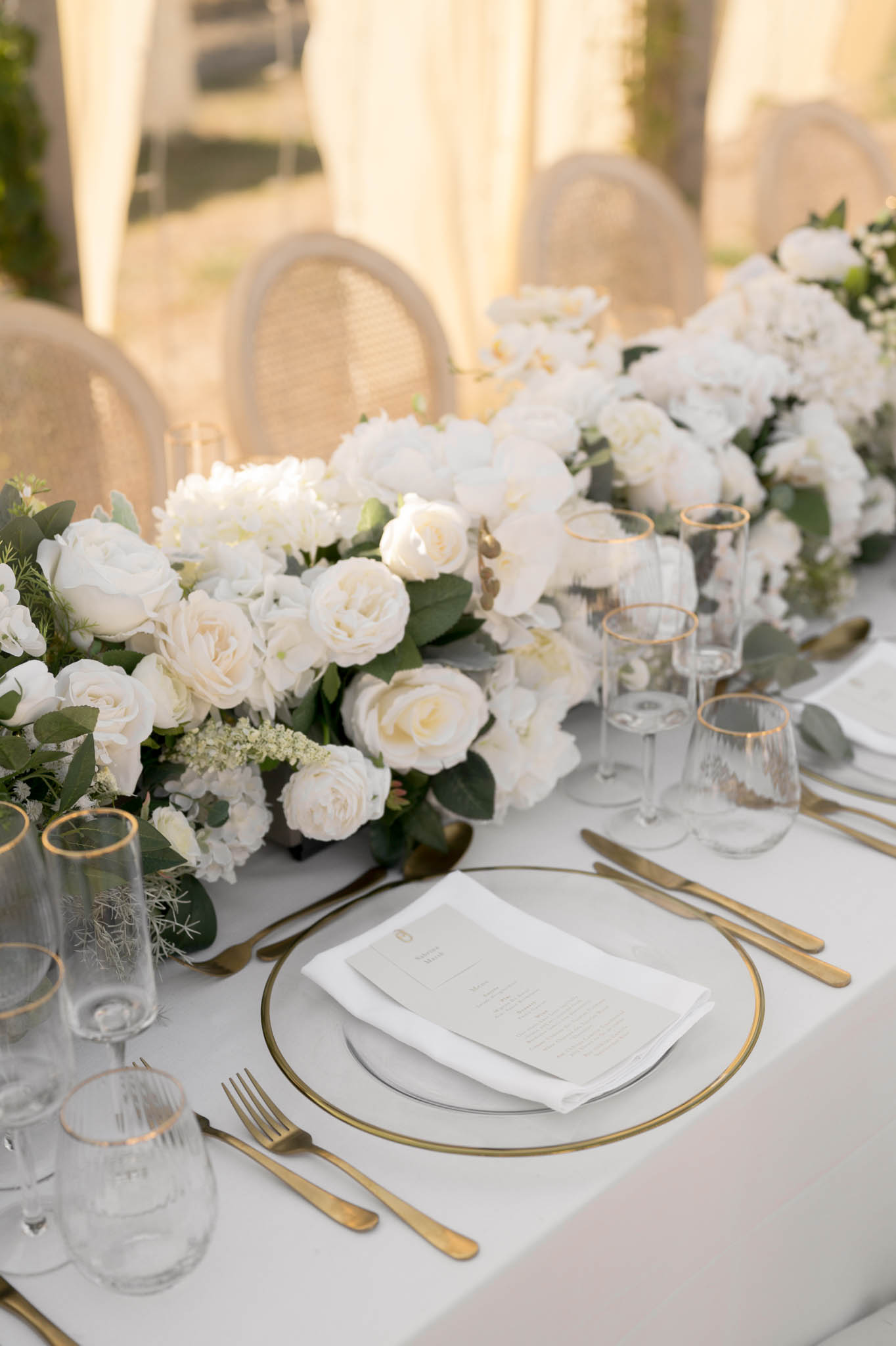Place setting with gold-rimmed glass charger, ivory roses and orchid runner, and rattan chairs