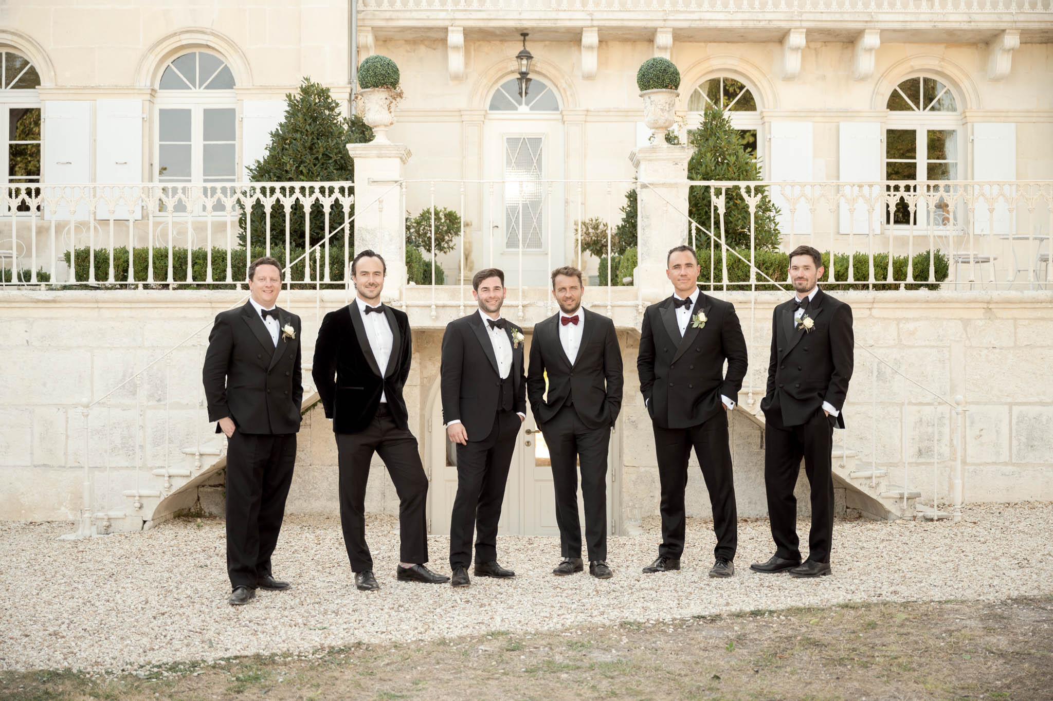 Six groomsmen in black tuxedos standing on gravel forecourt in front of cream limestone chateau with topiary