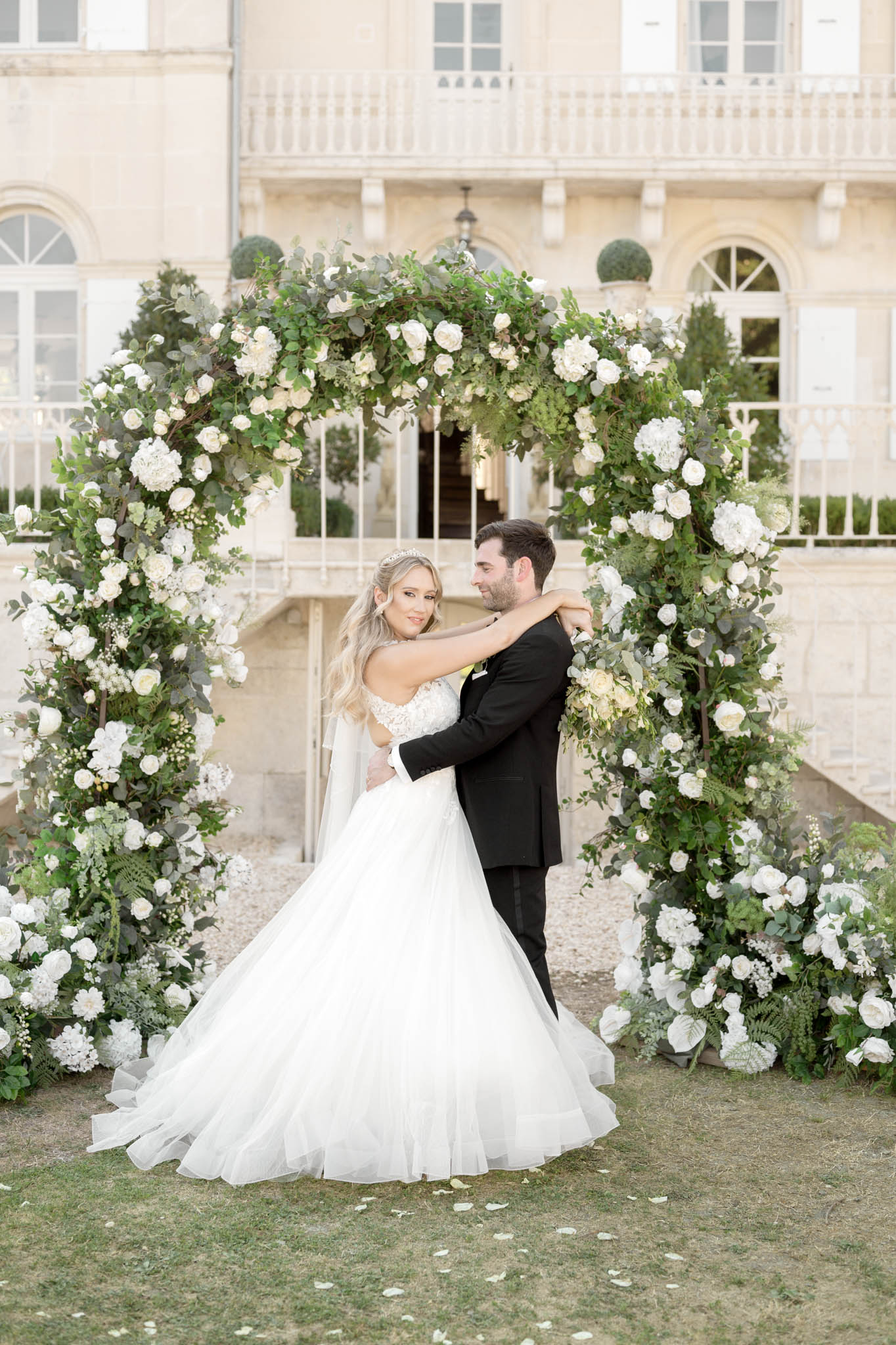 A couple portrait taken outdoors on the grounds of a French château, with the stone façade, arched windows, balcony, and ornamental topiary visible in the background. The bride and groom stand beneath a large circular floral arch densely decorated with white roses, white hydrangeas, and abundant greenery including eucalyptus, with matching floral clusters at the base of the arch and scattered white petals on the lawn. The bride wears a full ballgown with an off-the-shoulder lace bodice, cathedral-length veil, and a crystal tiara, while the groom wears a classic black tuxedo; the bride has her arm around the groom's neck as he holds her waist, with both looking at each other. The overall decor palette is white and green with a classic, formal styling aesthetic. Medium portrait shot framing the couple full-length within the arch.
