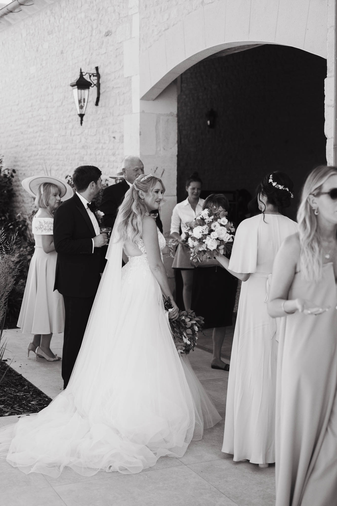 A black-and-white image of the bride and groom mingling with guests during what appears to be a cocktail hour or post-ceremony moment on an outdoor terrace. The setting features a stone building with an arched doorway and a wall-mounted lantern sconce. The bride wears a full ballgown with a lace bodice, a long cathedral-length veil, and a tiara hair accessory, with her hair in loose waves; she holds a large, full bouquet of roses and mixed foliage with dark-toned leaves. The groom is in a black tuxedo with a bow tie. Approximately six to eight guests are visible, including a bridesmaid in a long dress with flutter sleeves and a floral hair pin, and a female guest in a wide-brimmed hat and midi-length dress. The image is a candid mid-range shot with high contrast blacks and bright whites, capturing an informal interaction between the couple and their guests.