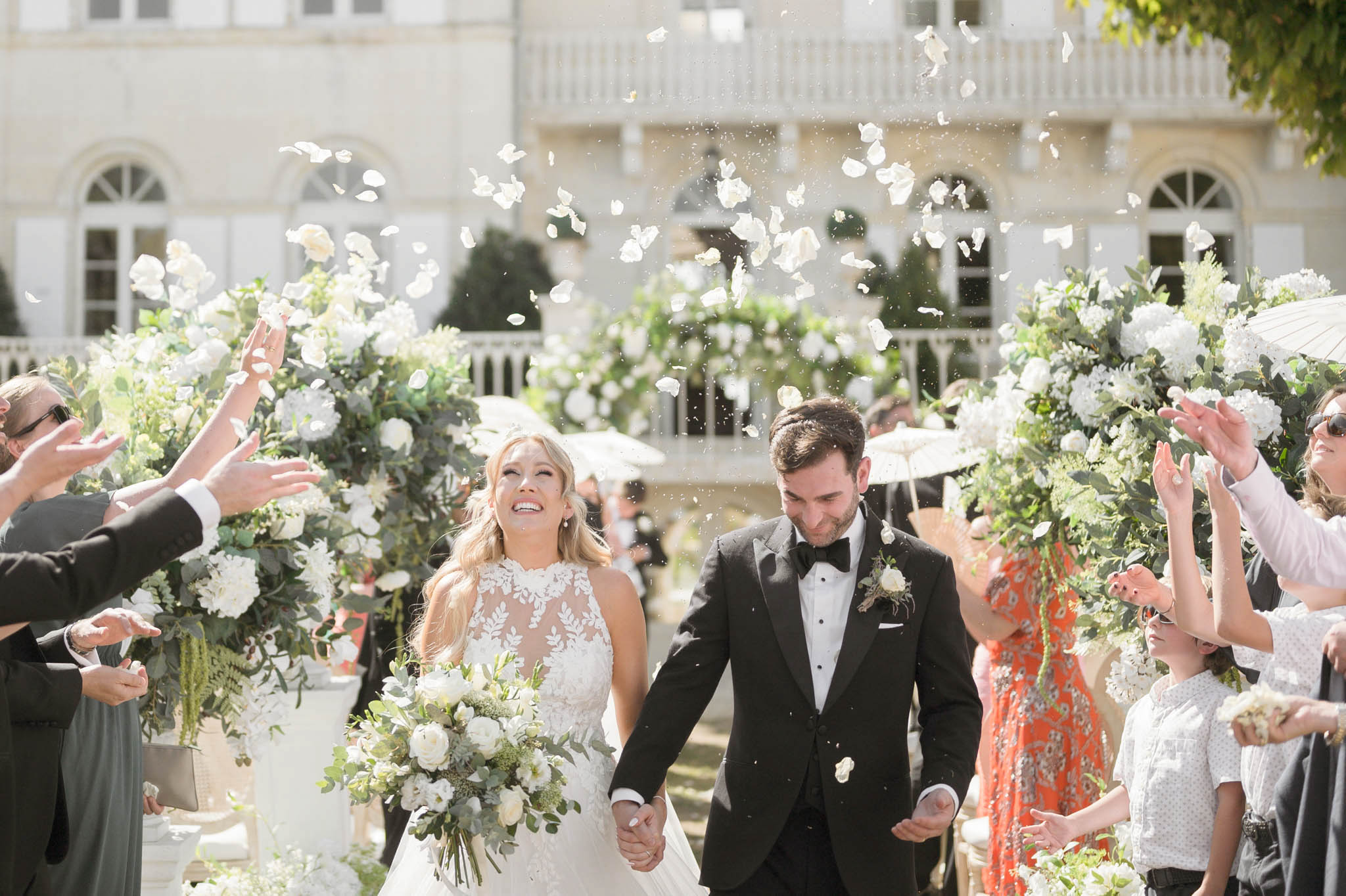 Couple recessional with rose petal toss past white hydrangea urns before chateau with arched windows