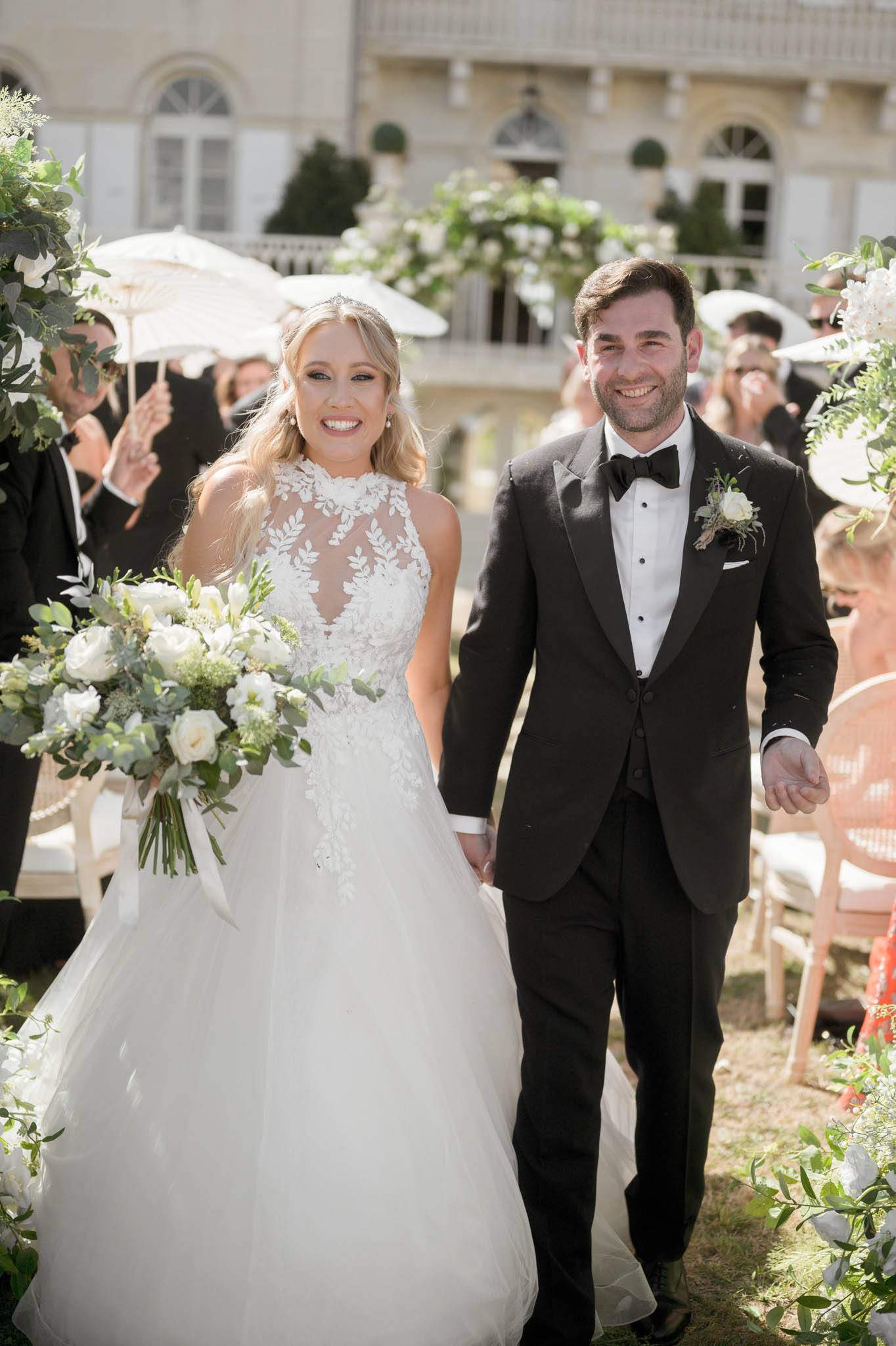 Bride and groom walk down the aisle after outdoor ceremony as guests cheer holding white parasols