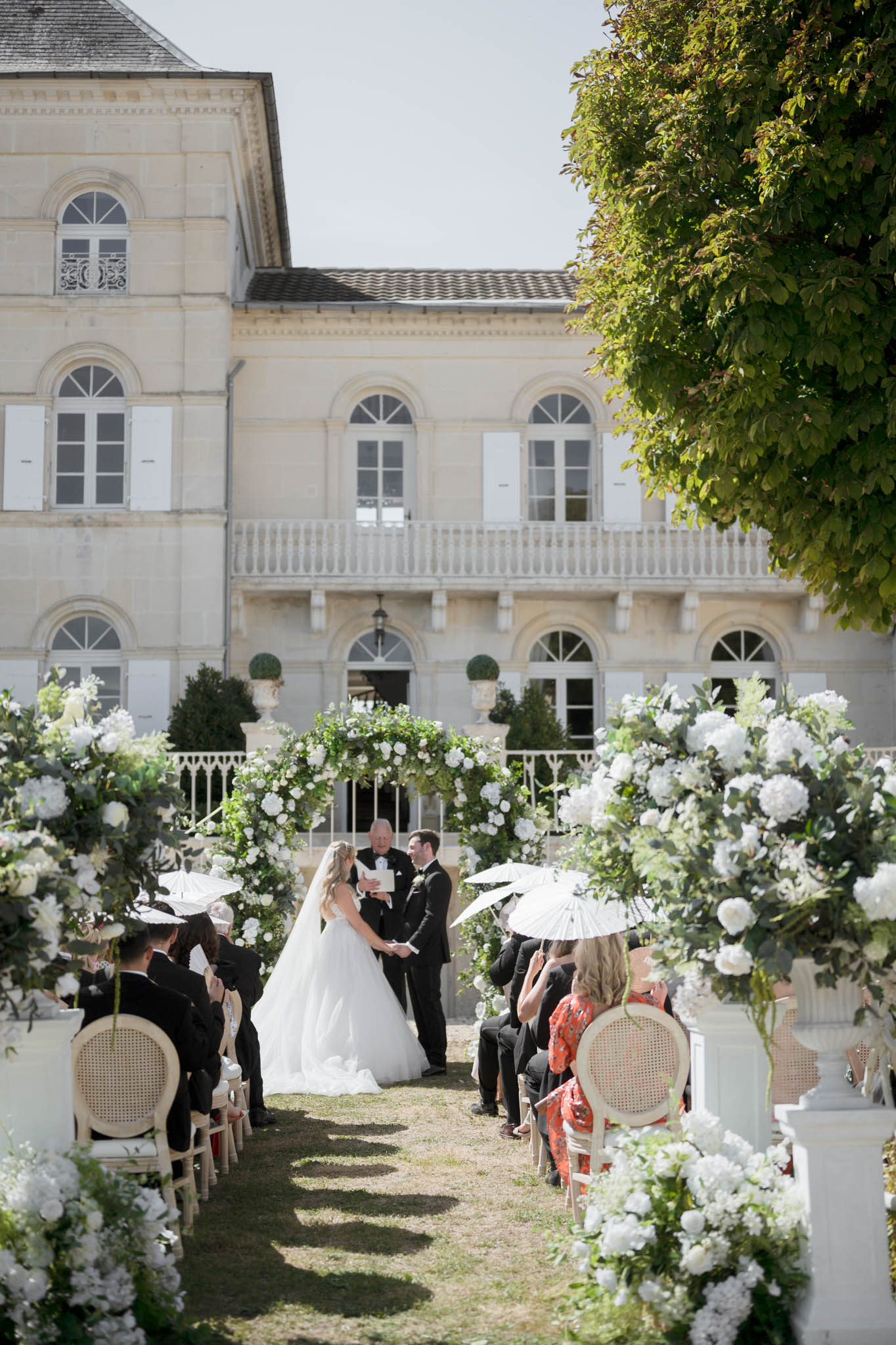 Outdoor ceremony with circular white rose arch guests on Louis XVI chairs and chateau facade backdrop