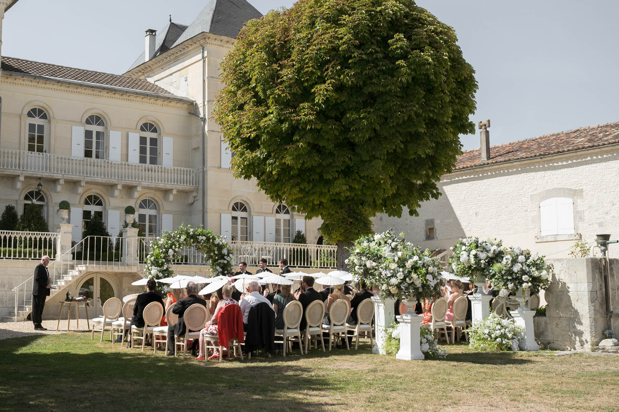 Ceremony courtyard with parasol-holding guests white rose arch and chateau balustrade staircase