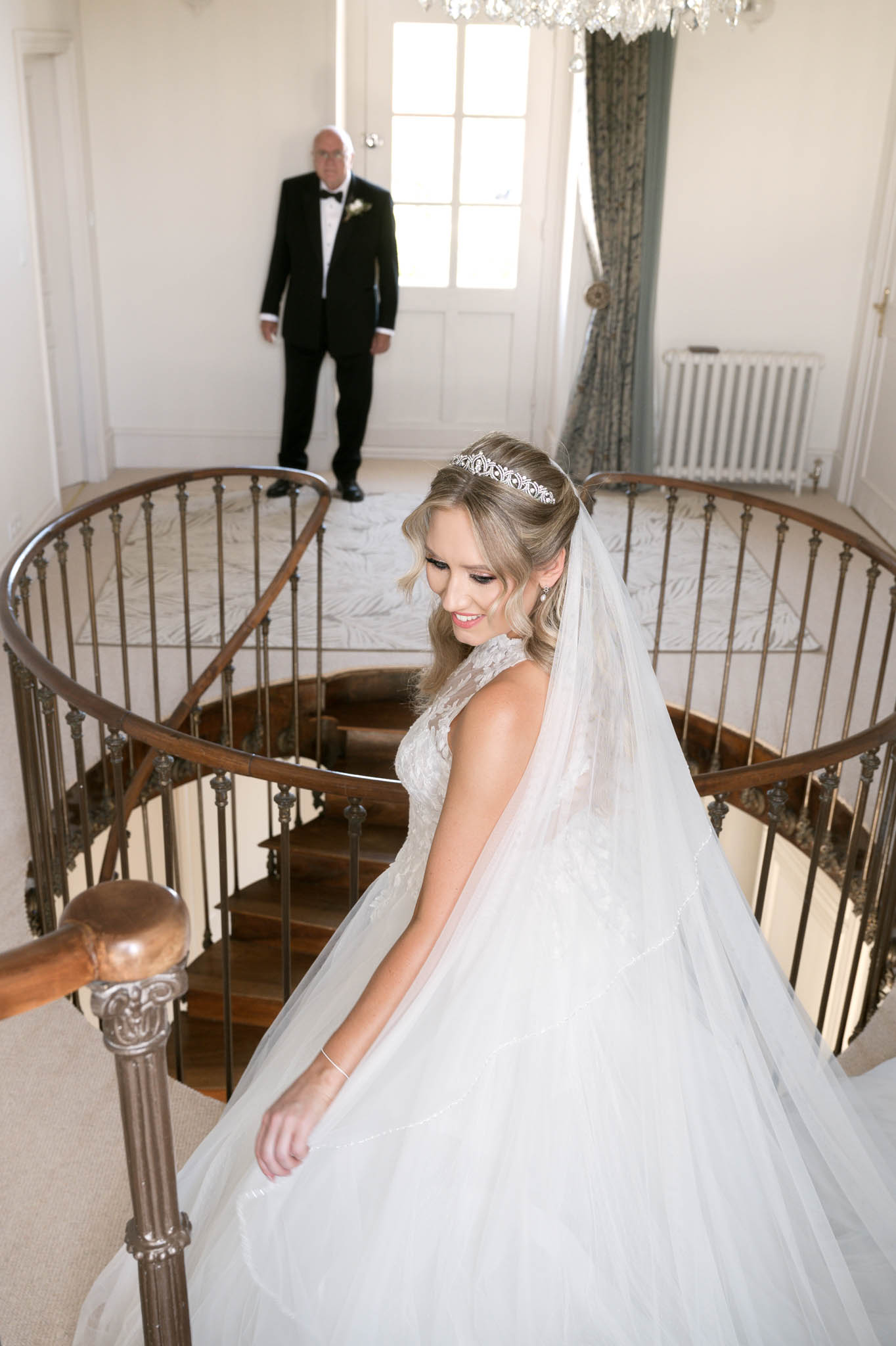 Bride in white ball gown and cathedral veil descending curved chateau staircase with father watching above