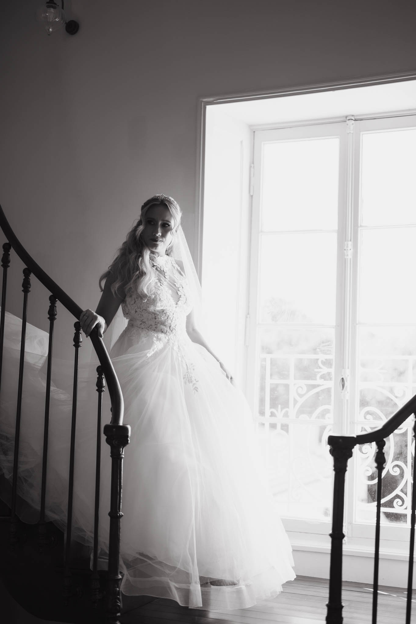 Bride in lace bodice tulle gown on curved chateau staircase with wrought-iron banister backlit in B&W