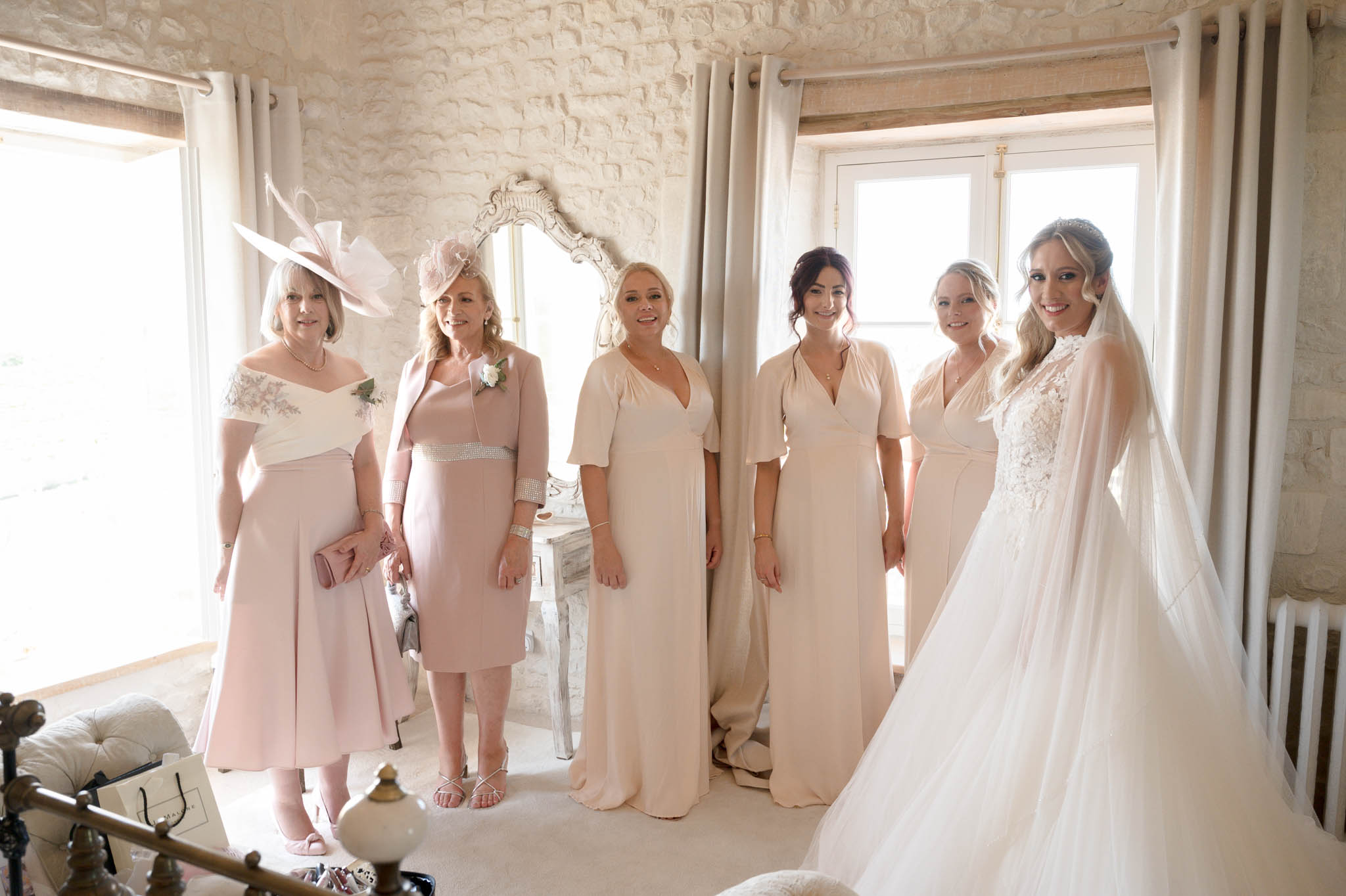 A getting-ready group portrait taken indoors in a bright, white-walled room with grey linen curtains, a ornate white vintage mirror on a dressing table, and a brass bed frame visible in the foreground. The bride stands on the right wearing a lace-bodice ballgown with a long tulle skirt and a cathedral-length veil, with her blonde hair styled up and a delicate hair accessory. She is joined by three bridesmaids in floor-length blush and champagne wrap-style dresses with short flutter sleeves, and two older women who appear to be mothers of the wedding party — one wearing a dusty pink midi dress with an off-shoulder lace top and a large blush fascinator hat, the other in a taupe-rose fitted dress with a rhinestone belt and a pink floral fascinator. The overall color palette is soft blush, champagne, and ivory throughout, giving the group a cohesive, classic look. The shot is a wide portrait taken at eye level with natural window light flooding in from behind the group.