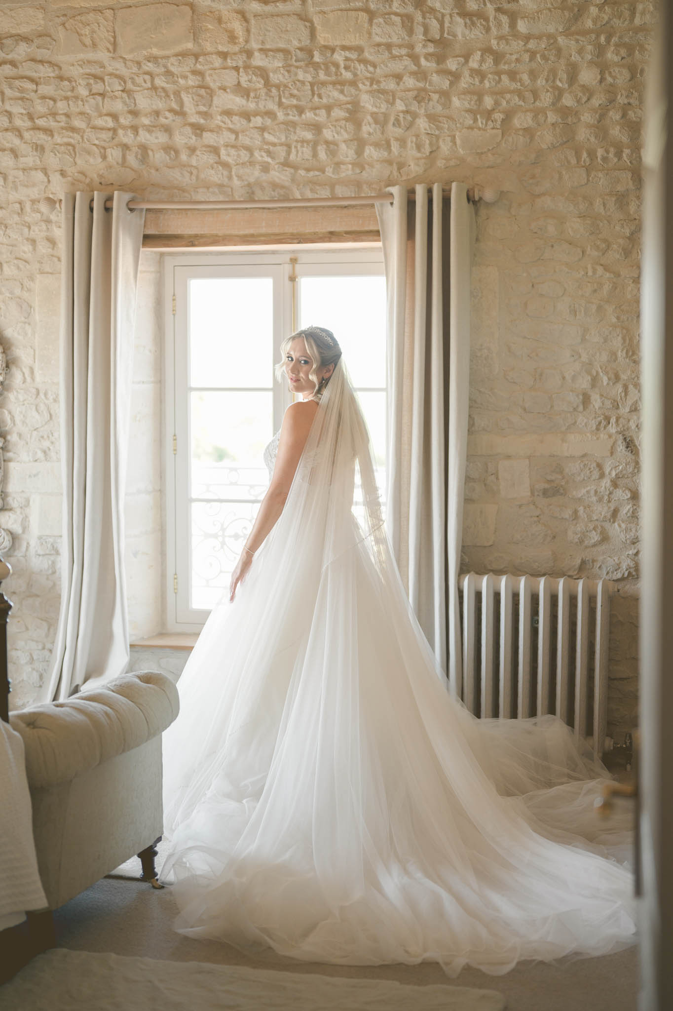 A bridal portrait taken indoors in a room with exposed limestone walls, likely within a French château or manor house. The bride stands three-quarter turned toward the camera in front of a tall window with natural backlight filtering through linen curtains, wearing a white full-ball-gown style dress with a voluminous tulle skirt and an extended cathedral-length train, paired with a long plain-edge veil and a crystal or pearl hairpiece at the crown of her upswept hair. She glances back over her shoulder toward the camera, with drop earrings visible. The room features a tufted cream settee to the left and a vintage-style white cast-iron radiator to the right, contributing to a classic French interior aesthetic. Full-length portrait composition with soft, warm window light.