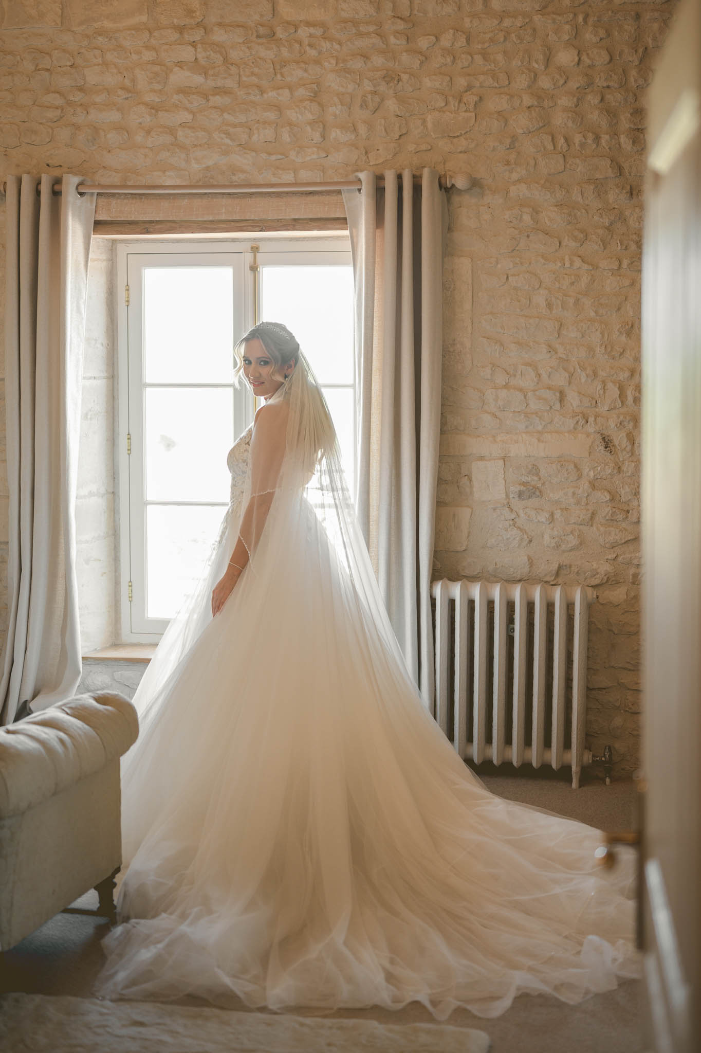 A bridal portrait taken indoors in a room with exposed limestone or pale stone walls, linen curtains, and a white cast-iron radiator. The bride stands in front of a tall French window with natural backlight, turning her head toward the camera with a smile. She wears a full-volume ivory tulle ballgown with a lace-bodiced corset back and a long cathedral-length veil with a delicate edge trim. Her hair is styled up with a small headpiece, and the gown's train is spread across the floor in front of her. A tufted cream sofa is partially visible on the left. The composition is a three-quarter portrait from behind, emphasizing the volume of the dress and veil against the bright window light.