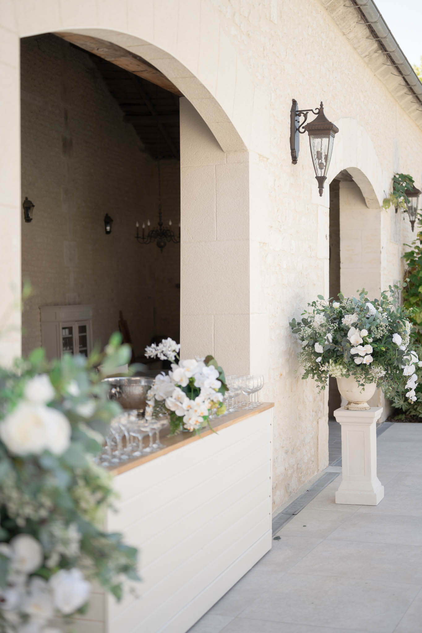 A cocktail hour setup photographed from outside a French château-style stone building, showing an outdoor bar service window cut into the pale limestone facade. The bar ledge is lined with coupe glasses, a silver champagne bucket, and arrangements of white orchids and white flowers with green foliage. A large white urn on a stone pedestal beside the arched doorway holds an abundant arrangement of white hydrangeas, white orchids, baby's breath, and eucalyptus. In the foreground, a second floral arrangement featuring white dahlias and eucalyptus is partially visible and slightly out of focus. Inside the arch, a dark iron chandelier is visible, along with additional wall-mounted lanterns on the exterior. The overall decor palette is white and green with classic styling. Wide environmental shot with no people present. Potential venue feature image.