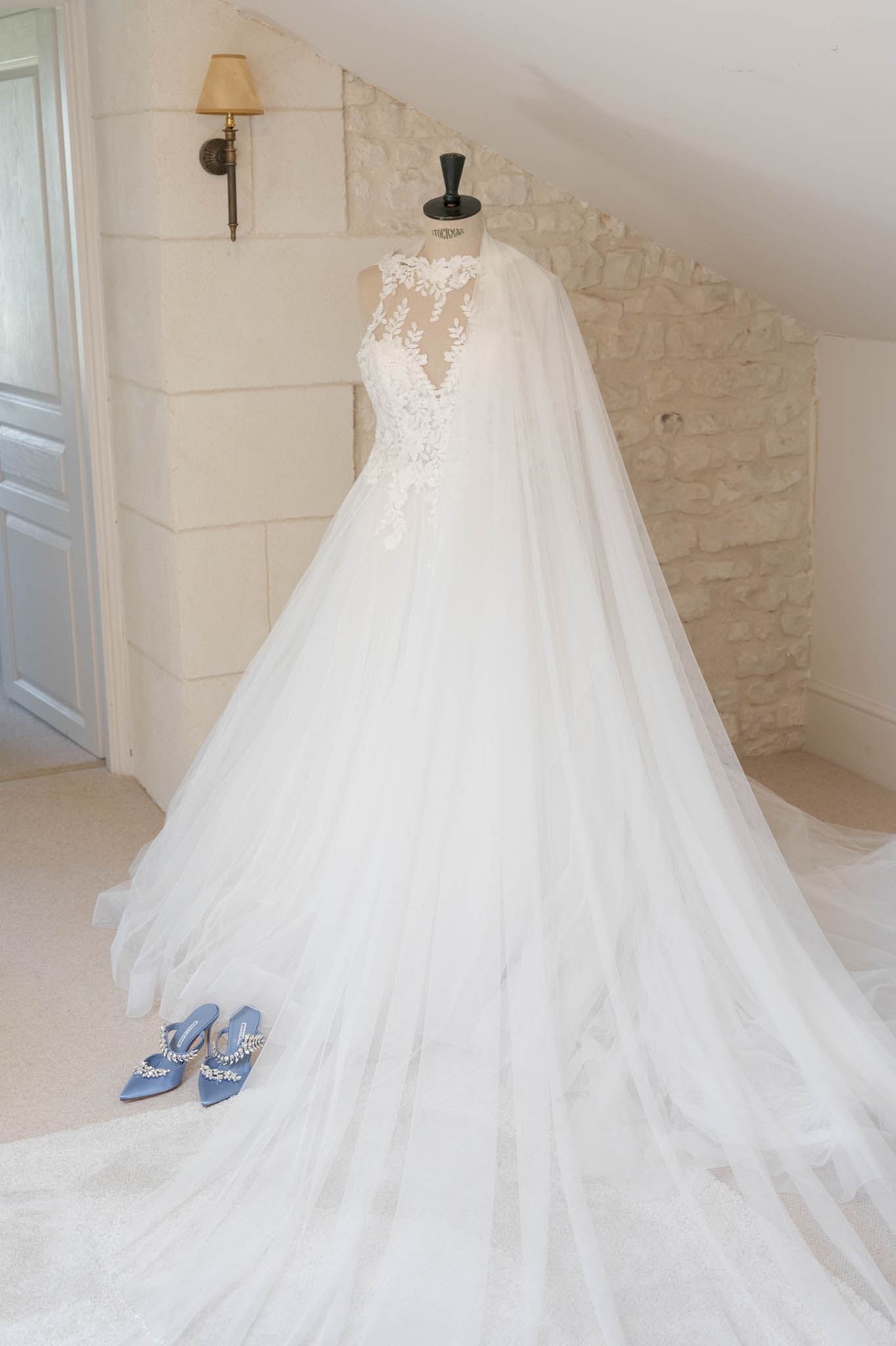 A getting-ready detail shot of a white bridal ball gown displayed on a dressmaker's mannequin in an indoor room with exposed limestone stone walls and a wall-mounted brass sconce lamp. The gown features an illusion lace bodice with floral and leaf appliqué, a deep V-neckline, and a voluminous layered tulle skirt with an extended cathedral-length train; a long sheer tulle cape or veil drapes from the shoulders. Placed at the base of the dress are a pair of powder blue satin pointed-toe mule heels adorned with crystal embellishments. The overall styling is classic and bridal, shot as a full-length wide portrait.