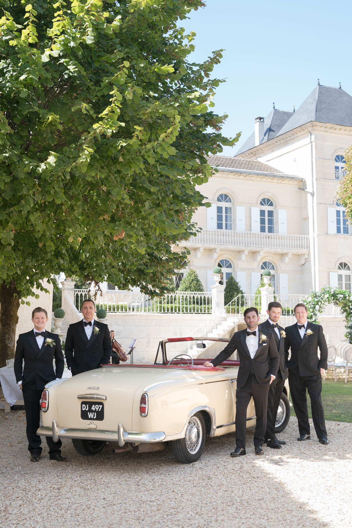 Five groomsmen in black tuxedos posing with a vintage Peugeot 403 convertible at a French chateau