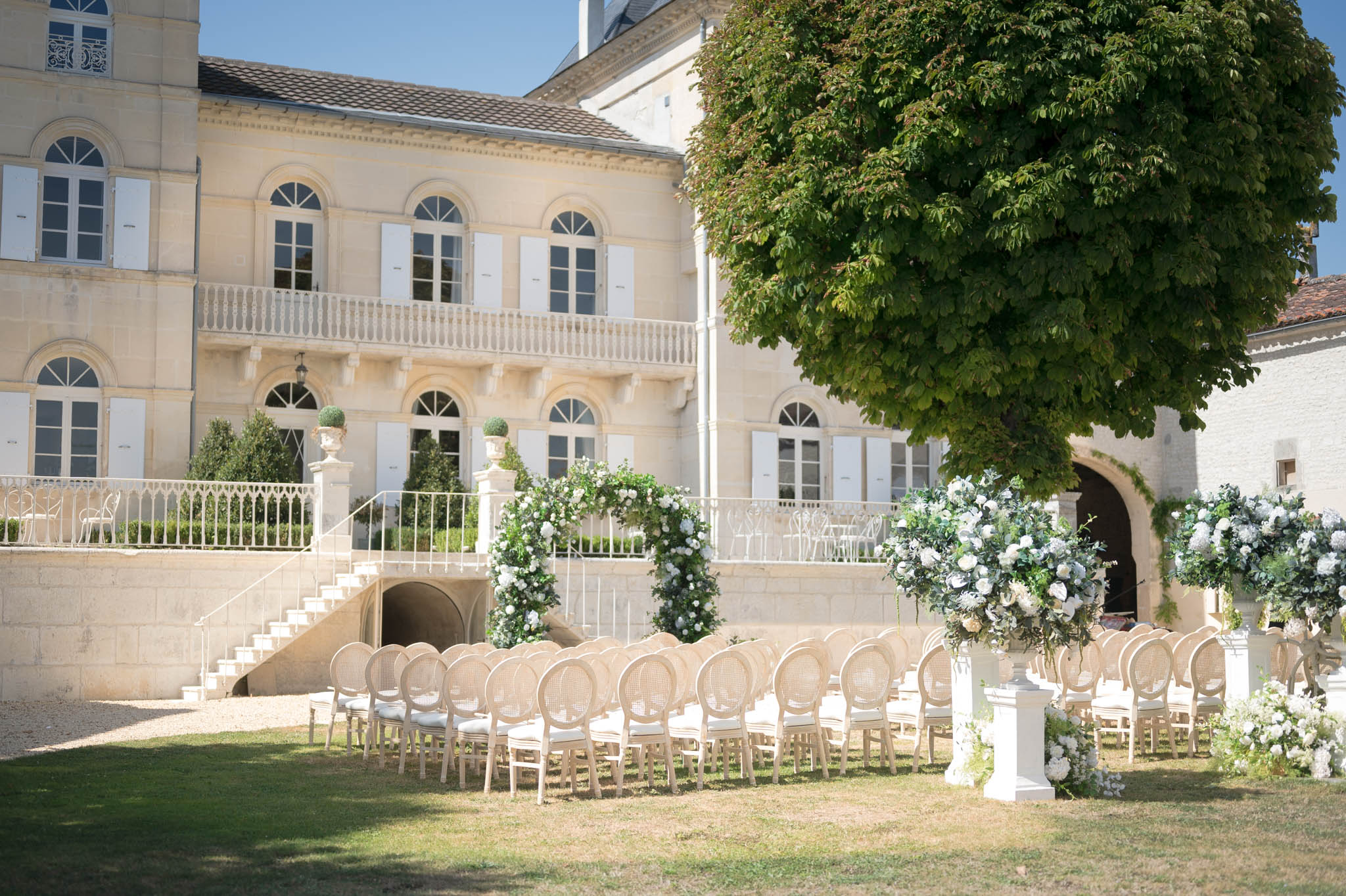 An outdoor wedding ceremony setup in the courtyard of a French château, with no guests or couple present — the space is arranged and ready for the ceremony. Rows of natural wood Louis XVI-style cane-back chairs with ivory cushions are arranged in two sections facing a floral arch positioned at the base of the château's stone staircase. The floral arch and large pedestal arrangements flanking the aisle feature white roses, white hydrangeas, and abundant green foliage. Additional oversized floral arrangements on white pedestals line the aisle on the right side. The décor palette is entirely white and green, creating a classic, restrained aesthetic. The wide shot captures the full ceremony layout against the backdrop of the château's cream limestone façade with white shutters, arched windows, and a wrought-iron balcony. Potential venue feature image.
