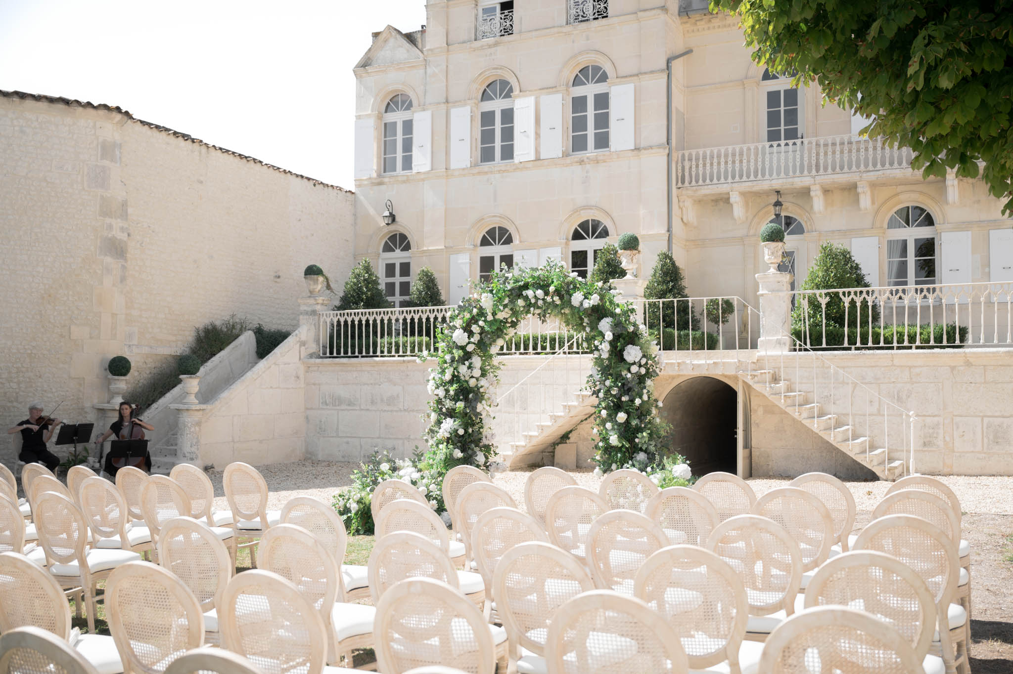 Ceremony setup with green and white floral arch, rattan Louis XVI chairs, and violin-cello duo at chateau