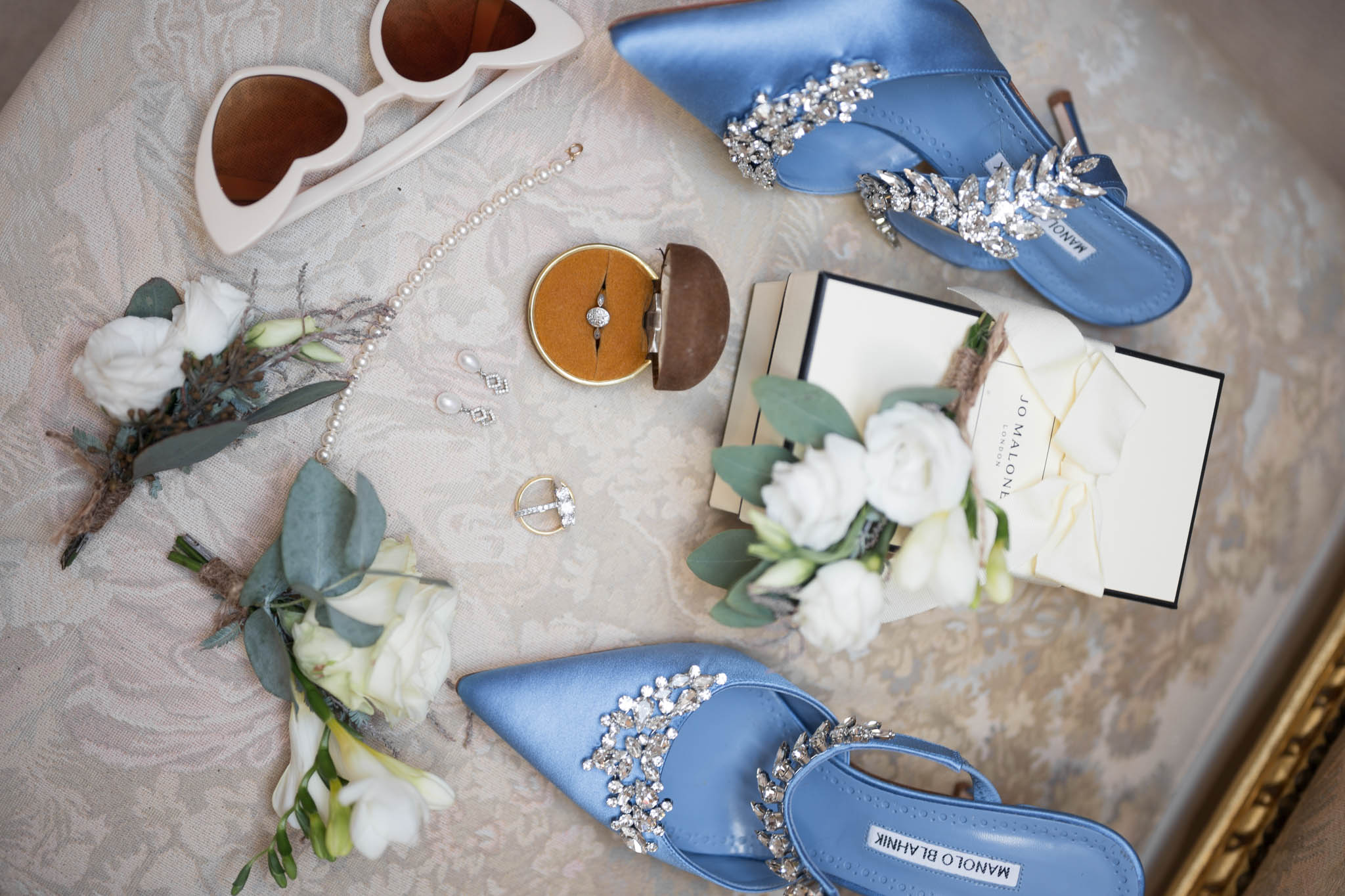 Flat lay of blue Manolo Blahnik shoes, pearl jewelry, diamond rings, and Jo Malone box on damask surface