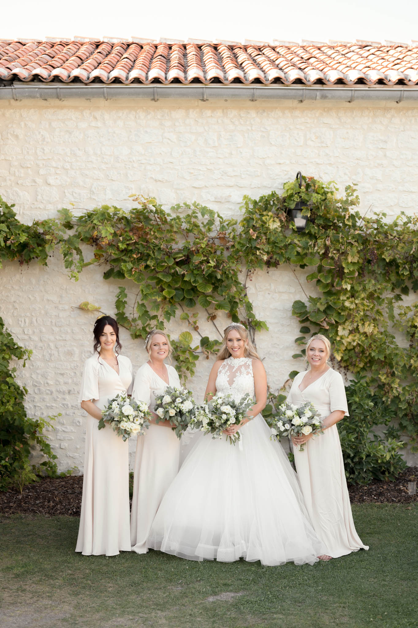 A bridal party portrait taken outdoors against a whitewashed stone building with a terracotta tile roof and climbing vines trained across the facade. The bride stands in the center wearing a full ballgown with a lace illusion high neckline and voluminous tulle skirt, accessorized with a delicate tiara. She is flanked by three bridesmaids in floor-length cream wrap-style dresses with flutter sleeves and V-necklines. All four women hold rounded bouquets composed of white roses, white ranunculus, and abundant eucalyptus and greenery foliage, maintaining a consistent white and green palette throughout. The styling is classic and cohesive, with a soft, monochromatic cream-and-white color scheme. This is a full-length group portrait shot in natural daylight.