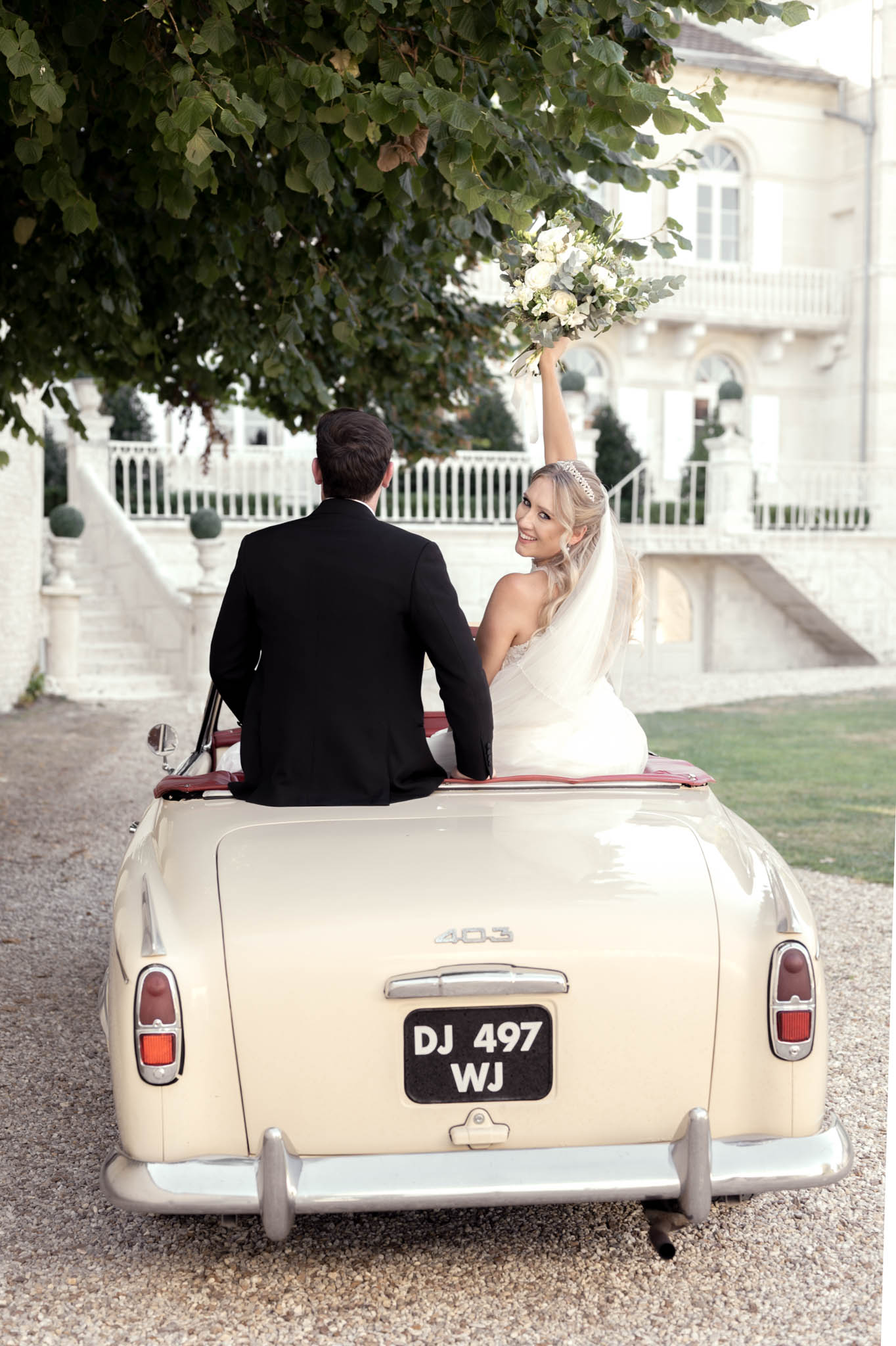 A couple portrait taken outdoors on a gravel driveway in front of a French château with white stone balustrades and steps. The bride and groom are seated in the open boot of a cream vintage Peugeot 403 convertible with a burgundy red interior. The groom wears a black suit and faces away from the camera, while the bride, wearing a fitted white sleeveless gown, a crystal tiara, and a long veil, turns toward the camera smiling with her arm raised holding a white and green bouquet of roses and eucalyptus. The bouquet is lifted high above her head in a celebratory gesture. The shot is taken from behind the car at mid-distance, framing the couple against the château facade.