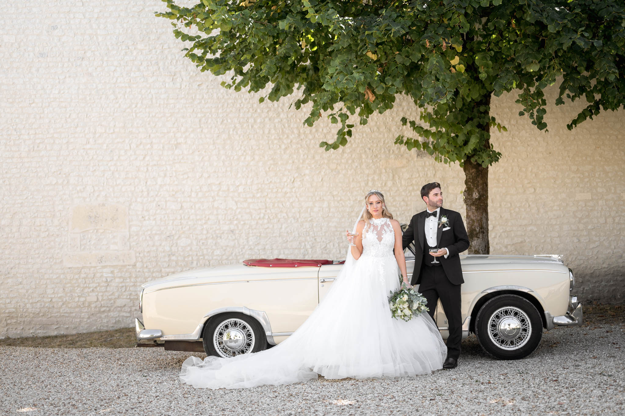 Couple leaning on vintage cream convertible with red interior bride in illusion lace ballgown with tiara groom in tuxedo