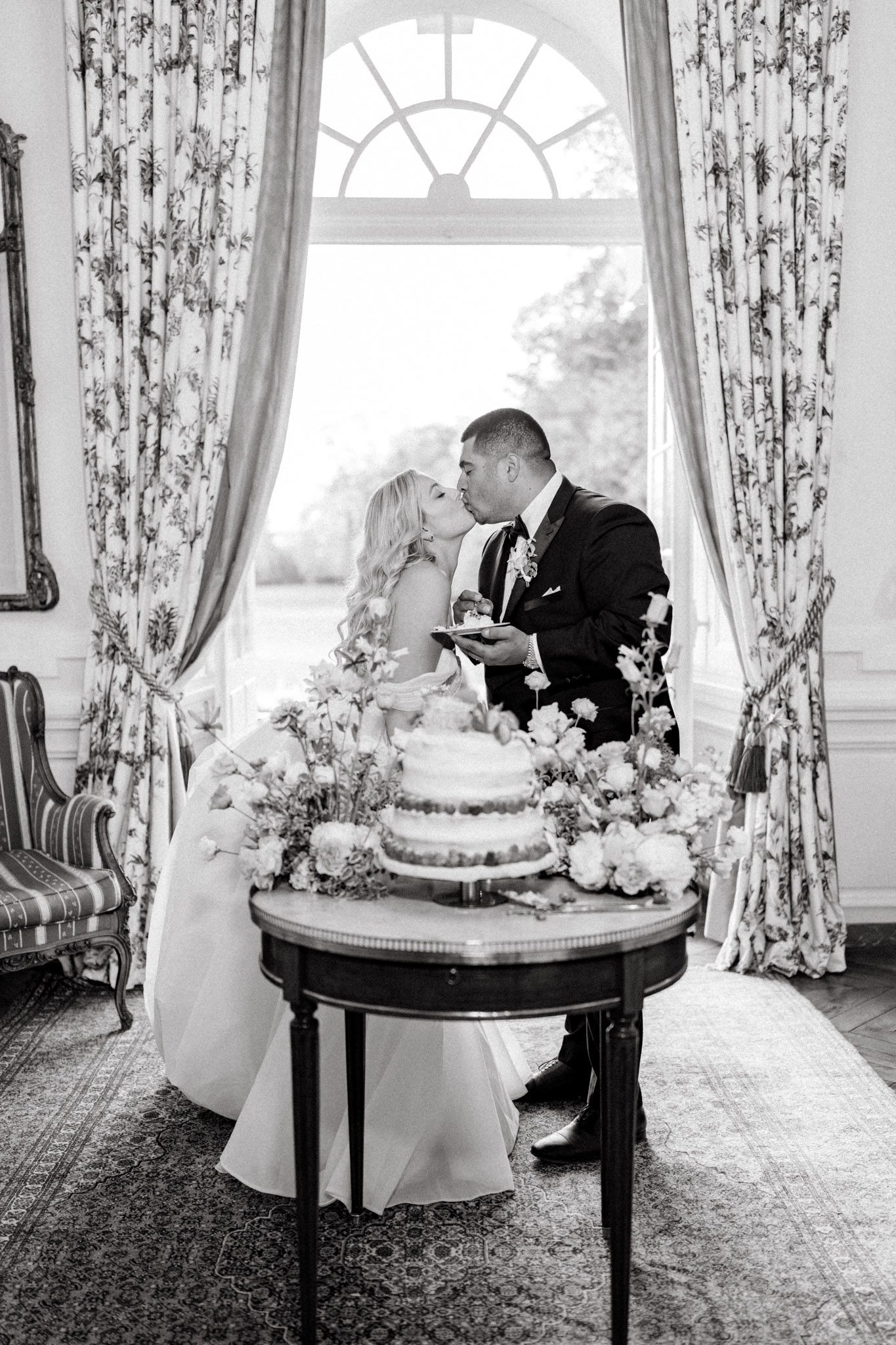 Black-and-white image of a bride and groom sharing a kiss during the cake cutting moment inside a formal reception room. The bride wears a strapless fitted gown with a full skirt and has long wavy hair; the groom is dressed in a dark tuxedo with a boutonniere and holds a plate with a slice of cake. The three-tiered wedding cake sits on a small dark wood side table and is surrounded by loose floral arrangements featuring peonies, ranunculus, and delicate branching blooms. The room features floor-to-ceiling floral-print curtains with tassel tiebacks, wood-paneled walls, a patterned area rug, and a large arched window behind the couple that floods the scene with light. A striped upholstered armchair and an ornate framed mirror are visible to the left, suggesting a classic French château interior. The composition is a medium-wide portrait shot centered on the couple with the architectural window framing them from behind.