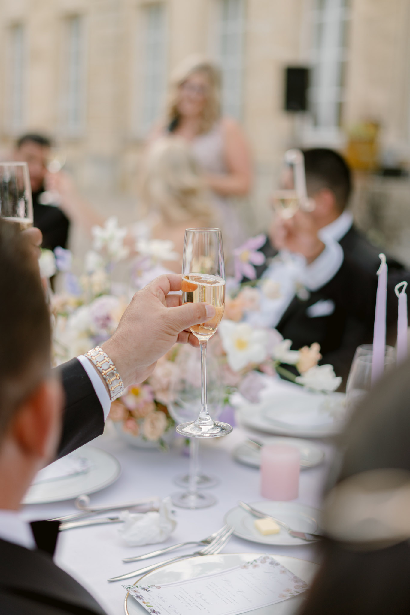 A champagne toast during an outdoor wedding reception, with a guest in the foreground raising a flute of rosé champagne toward the camera. The table is dressed in a white linen and features low floral centerpieces in a soft palette of blush, peach, lavender, and white — including tulips and open garden roses — alongside tall lavender taper candles and pink votive candles. White plates, silver cutlery, and printed menus are visible at each place setting. In the background, a woman in a blush or light grey dress stands holding a microphone, appearing to give a speech, while other guests in formal black tie attire raise their glasses. The setting is an outdoor courtyard in front of a classical stone building with tall windows. The shot is taken from a seated guest's perspective, with shallow depth of field keeping the foreground glass sharp and the background softly blurred.