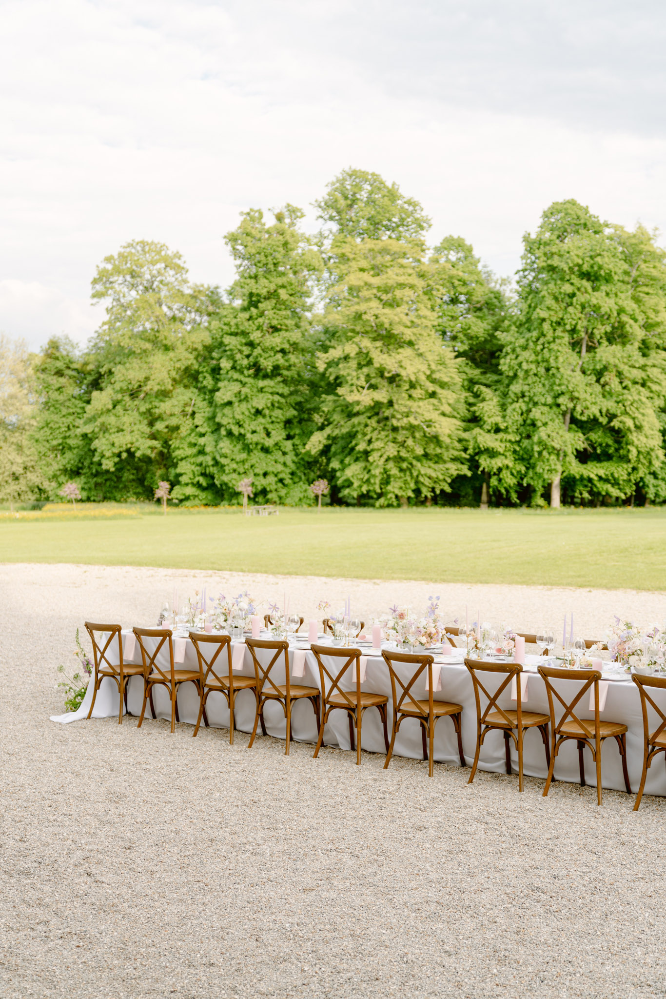 An outdoor wedding reception table setup photographed in a wide shot on a gravel surface, with an open lawn and mature trees in the background. The long rectangular table is dressed in a light grey linen and lined on one visible side with approximately ten warm-toned wooden cross-back chairs. The tablescape features low floral arrangements in blush pink, lavender, and white tones with loose, garden-style blooms, paired with tall dusty pink taper candles, clear glassware, and grey or sage-toned place settings. Pink napkins are folded at each place setting, contributing to a soft, muted pastel palette. The overall styling is classic French countryside with a romantic, airy aesthetic.