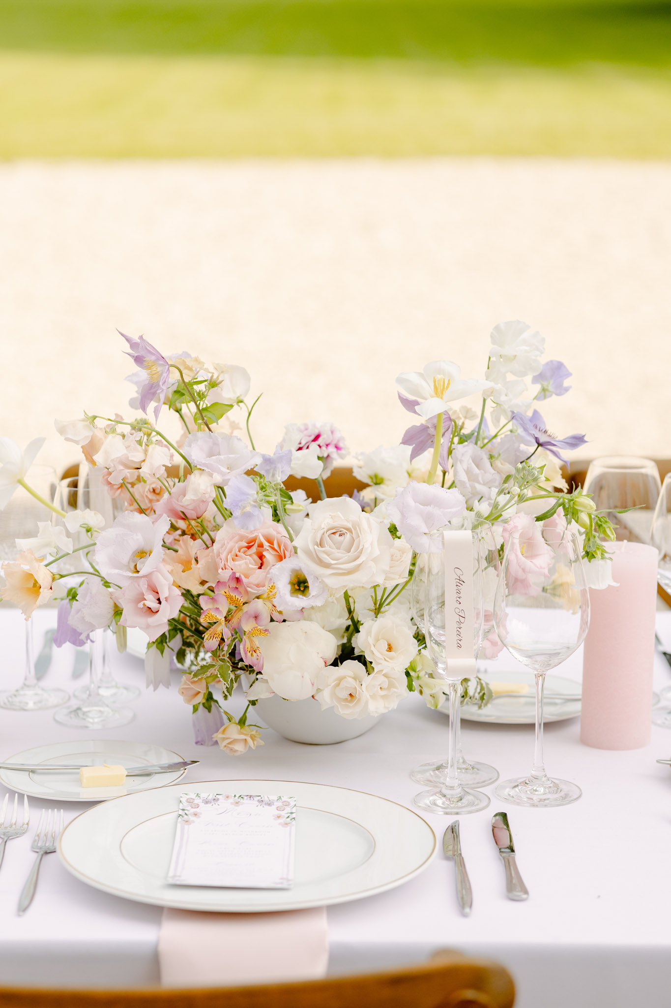 A close-up detail shot of an outdoor wedding reception table setting. The centerpiece consists of low, lush floral arrangements in a white bowl featuring blush and peach garden roses, white ranunculus, pale lavender lisianthus, lilac columbine, sweet peas, alstroemeria, and trailing green foliage. The table is dressed in a white linen cloth with a pale blush pink napkin at the place setting. Tableware includes a charger plate with a thin gold rim, a white dinner plate topped with a floral-illustrated menu card, and silver cutlery. A wine glass is personalized with hand-lettered calligraphy in the name 'Marie Preva,' and a blush pink pillar candle sits to the right. The overall decor palette is soft pastel — blush, white, peach, and lavender — with a romantic garden style. Mustard-yellow upholstered chairs are partially visible in the background.