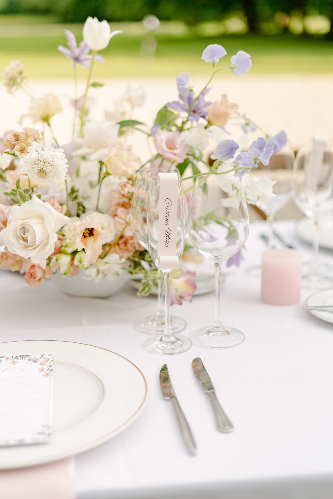 A close-up detail shot of an outdoor wedding reception table setting. The table is dressed in a white linen cloth and features a lush floral centerpiece composed of ivory roses, blush carnations, white scabiosa, lavender sweet peas, white tulips, and soft peach lisianthus arranged in a low white vessel. In the foreground, a champagne flute holds a cream ribbon place card with calligraphy script reading 'Cristina Mills,' positioned alongside a wine glass. The place setting includes white plates with a thin gold rim, silver flatware, a floral-printed menu card, and a small dusty pink pillar candle. The overall decor palette is soft pastel — blush, ivory, lavender, and peach — with a romantic garden aesthetic.