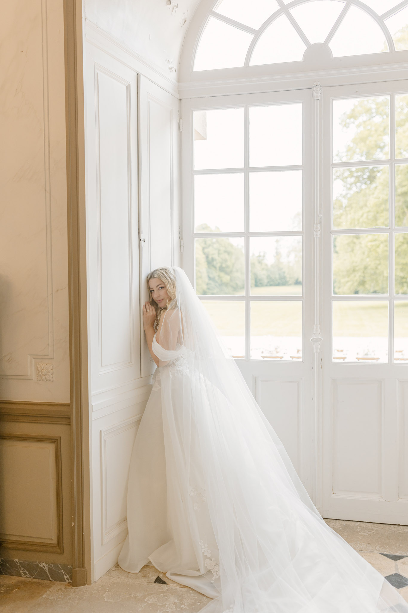 A bridal portrait taken indoors at a French château, featuring the bride standing and leaning against white-painted wood-panelled walls beside a large arched window with floor-to-ceiling white-framed panes. The bride wears an ivory tulle ballgown with a low open back, thin spaghetti straps, a beaded waistband detail, and a long cathedral-length veil; the full skirt and veil trail across the marble-tiled floor. She looks back over her shoulder toward the camera with a relaxed pose. The room features classic French interior architecture including warm cream-toned wainscoting, ornate door frames, and a decorative rosette panel. Natural light floods in through the arched window. The composition is a full-length portrait shot.