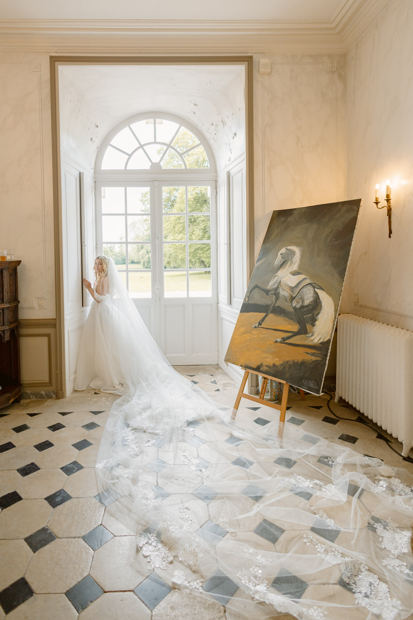 Bride with cathedral lace veil spread across geometric tile floor gazing through arched French doors in chateau