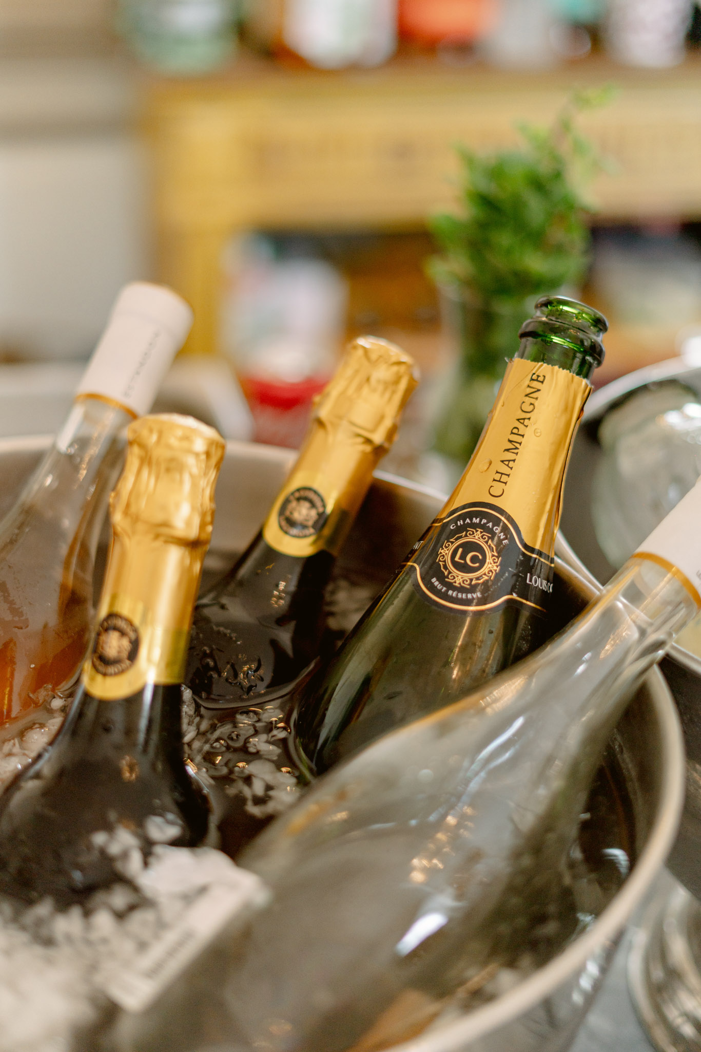 A close-up detail shot of a silver ice bucket containing multiple champagne and wine bottles chilling for a wedding reception or cocktail hour. The most prominent bottle is a Champagne LC (Loursac) Brut Réserve with a black and gold label and green glass, flanked by two additional bottles with gold foil necks. A fourth clear glass bottle, likely still wine, is also visible in the bucket. The background is softly blurred but shows what appears to be an indoor bar or service area with shelving and a small green herb garnish. The image is shot at a shallow depth of field, keeping the bottle labels in sharp focus.