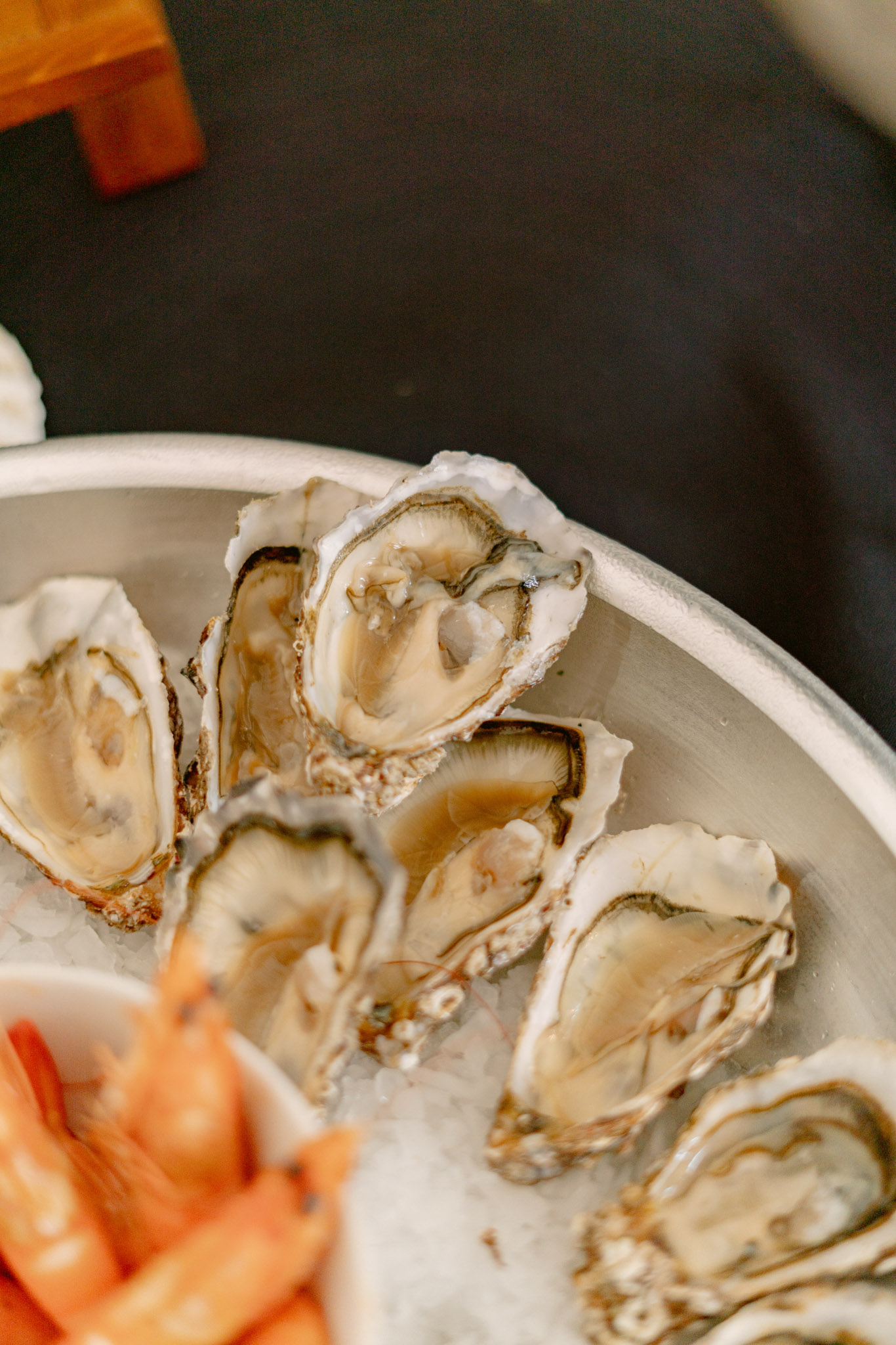 Close-up detail shot of a catering or cocktail hour food display featuring freshly shucked oysters arranged on crushed ice in a round silver metal tray. Approximately seven to eight open oyster half-shells are visible, showing pale cream and grey-toned flesh. In the lower left corner, orange-pink cooked shrimp are partially visible, suggesting a seafood platter presentation. A small wooden block or stand is partially visible in the upper left background. The setting appears to be indoors against a dark surface, with natural or ambient soft lighting. The composition is a tight overhead angle focusing on the food detail.
