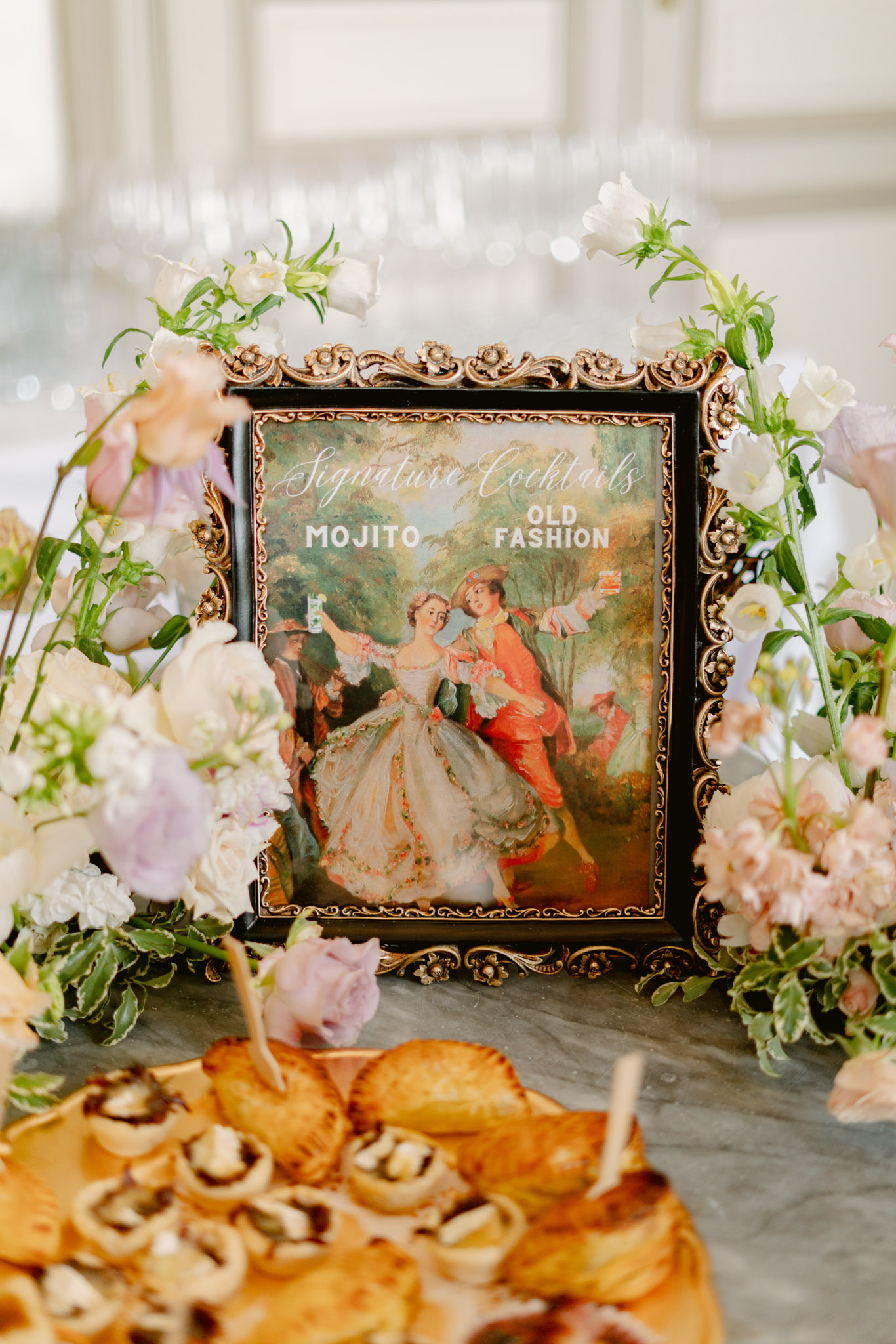 Signature cocktail sign in ornate gold frame surrounded by blush and lavender flowers with canape platter